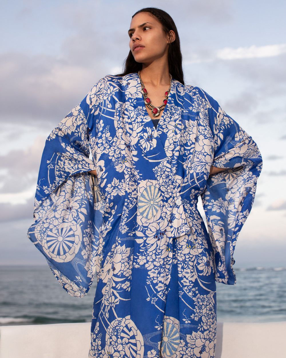 A woman stands outdoors by the ocean, wearing the Grace Royal Spoke Kimono Dress with a blue flower cart motif and a red beaded necklace, gazing to the side under a cloudy sky.