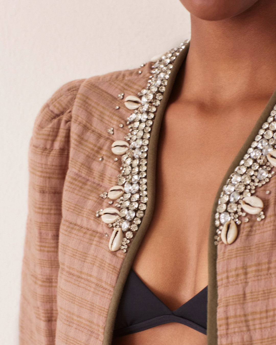 Close-up of a person wearing the Pasha Shell and Gem Jacket, a brown hand-embroidered piece featuring rhinestones and cowrie shells along the collar. The jacket is slightly open, revealing a black top underneath against a neutral beige wall backdrop.