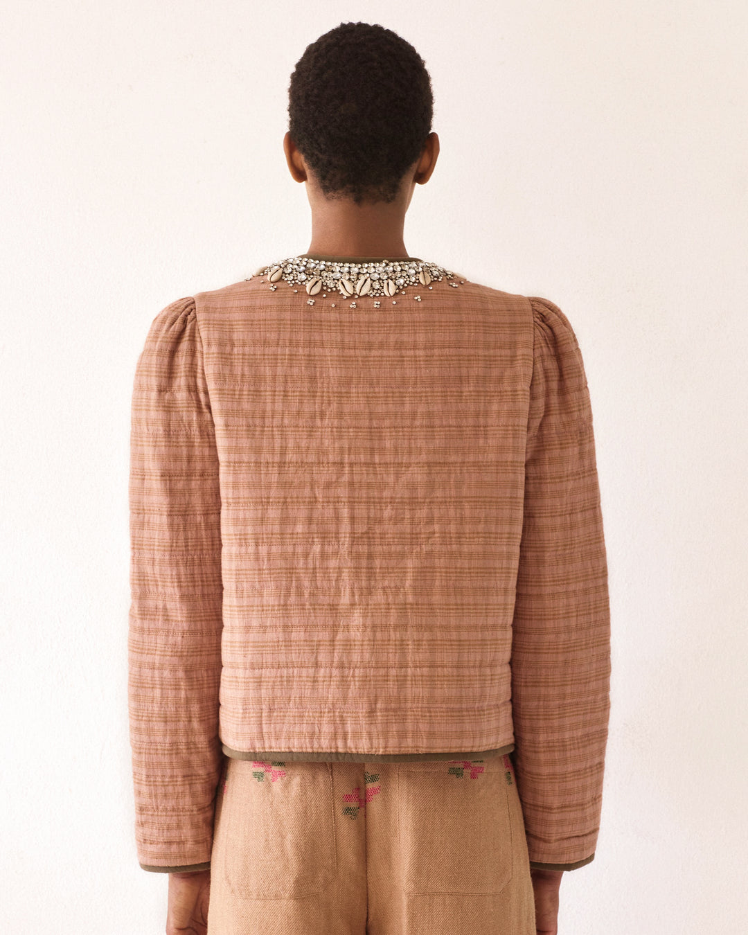 A back view of a person wearing the Pasha Shell and Gem Jacket, which features a textured brown ikat pattern with puffed sleeves, paired with a beaded necklace adorned with cowrie shells. The jacket is matched with tan pants showcasing subtle floral embroidery against a plain, light-colored wall backdrop.