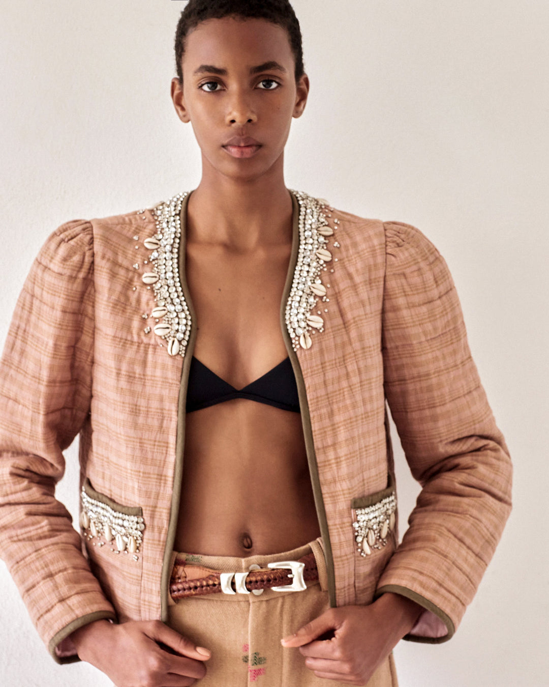 A person with short hair is wearing a stylish Pasha Shell and Gem Jacket, which features intricate beadwork around the collar. Underneath, they wear a black crop top and beige trousers, standing against a plain background.