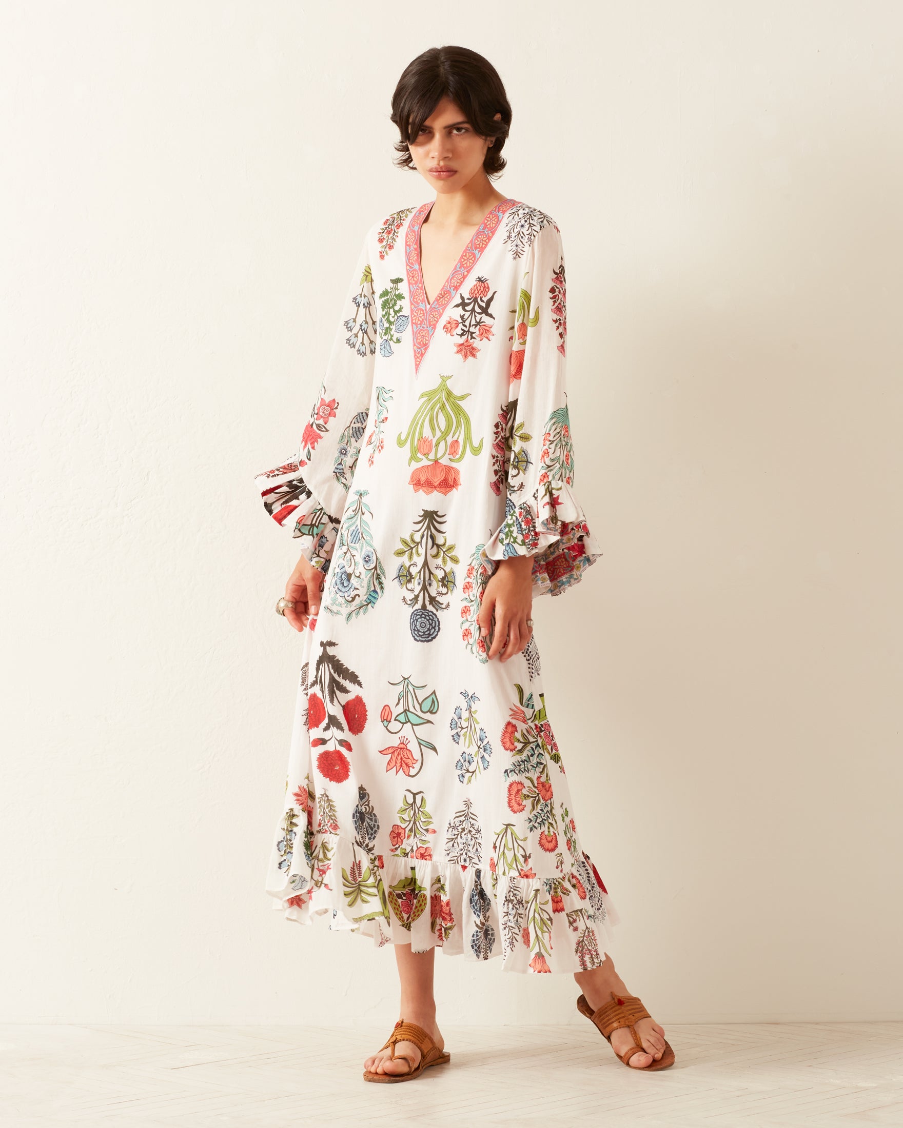 Against a plain wall, an individual poses in the eye-catching Zelda Flower Show Dress, adorned with vibrant floral patterns. This hand-printed cotton dress showcases a pink V-neckline along with charming ruffled sleeves and hem. They complete the look with brown sandals and have short, dark hair.