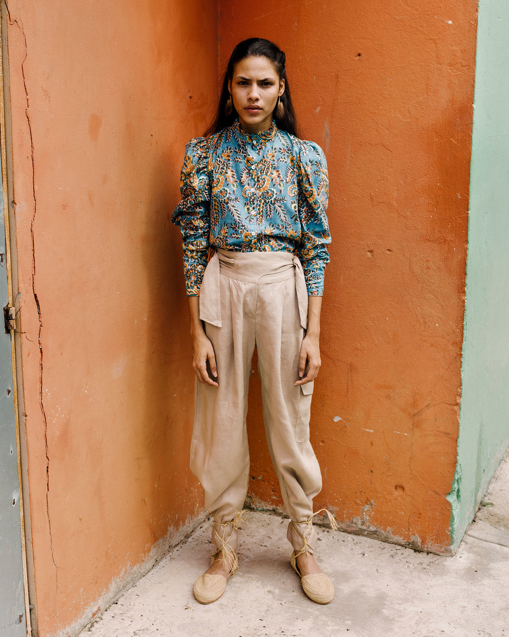 A woman stands against a two-toned wall, showcasing a patterned blouse, Painter Sand Linen Pants, and lace-up shoes. The background is painted in vibrant orange and green hues. Her long hair cascades softly as she maintains a neutral expression.