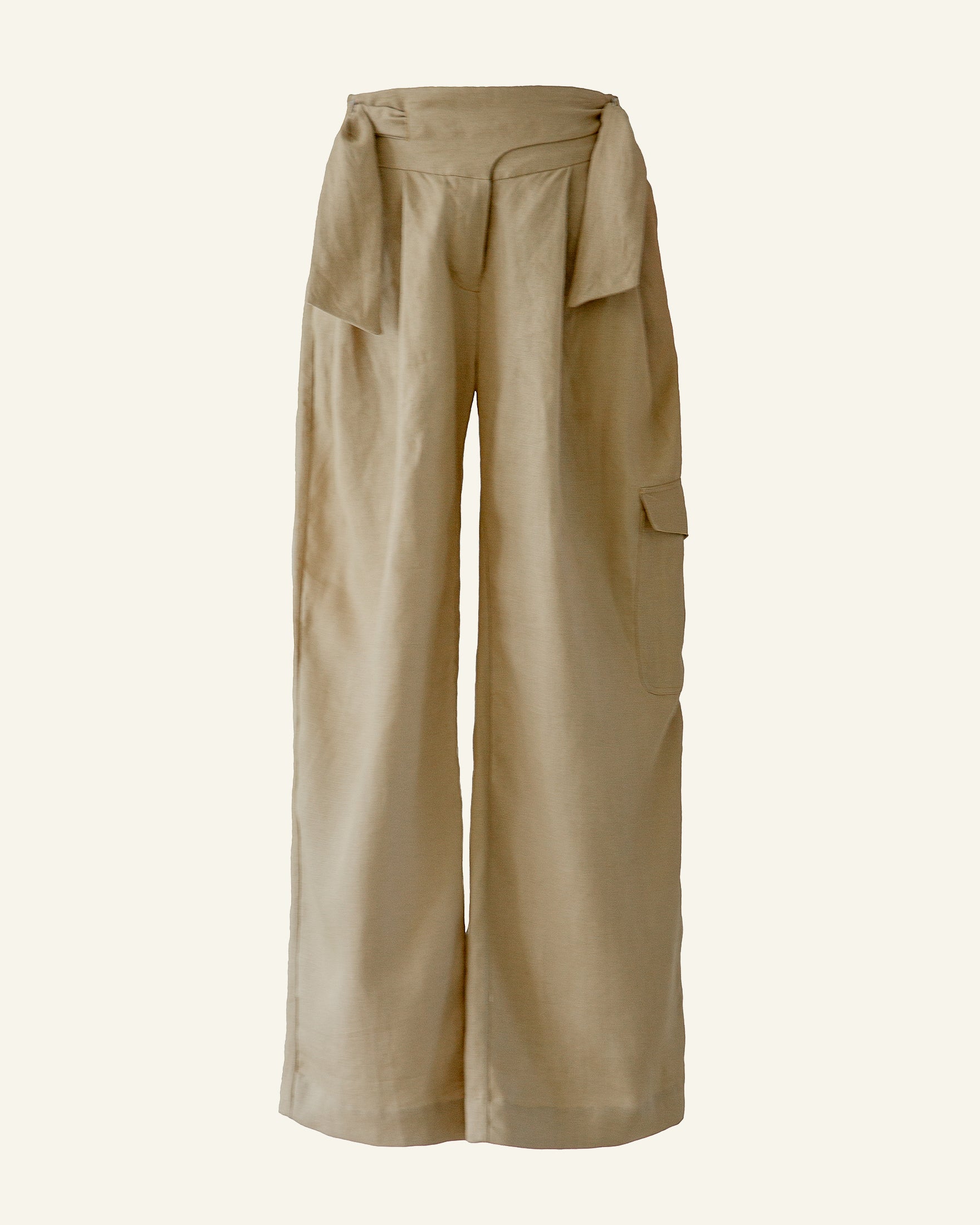 The Painter Sand Linen Pant is a pair of high-waisted, wide-legged beige women's trousers made from cotton. They feature a draped fabric detail at the waist and a cargo pocket on the right leg, showcasing a minimalist design against a plain background.