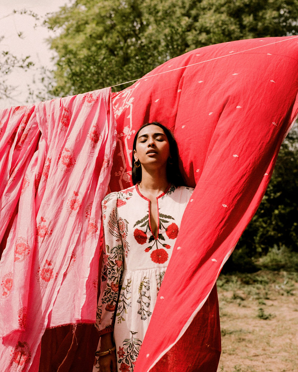 A woman stands outdoors beside vibrant red and pink garments hanging on a clothesline. She wears a Winslow Flower Show Dress, crafted from hand-printed cotton with red floral patterns, set against a background of greenery and open sky.