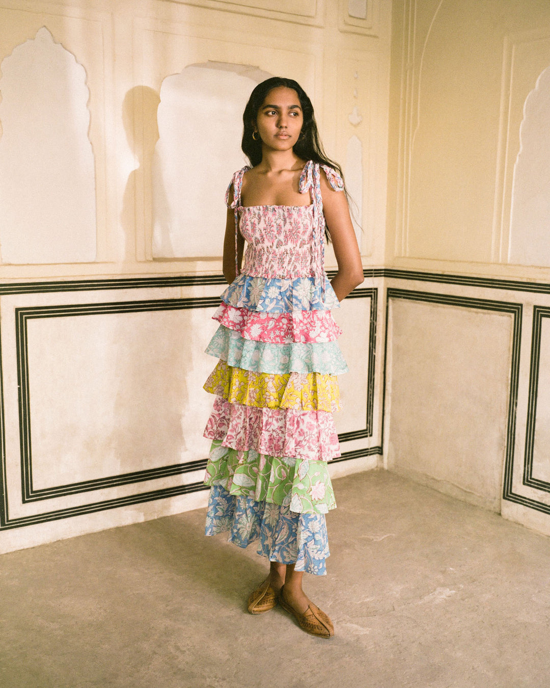 A woman stands in a room with ornate walls, wearing the Club Tropical Ruffle Dress adorned with vibrant patterns and colors—pink, blue, green, and yellow. Its smocked bodice adds elegance. She pairs it with brown woven shoes and has long, dark hair cascading down her shoulders.