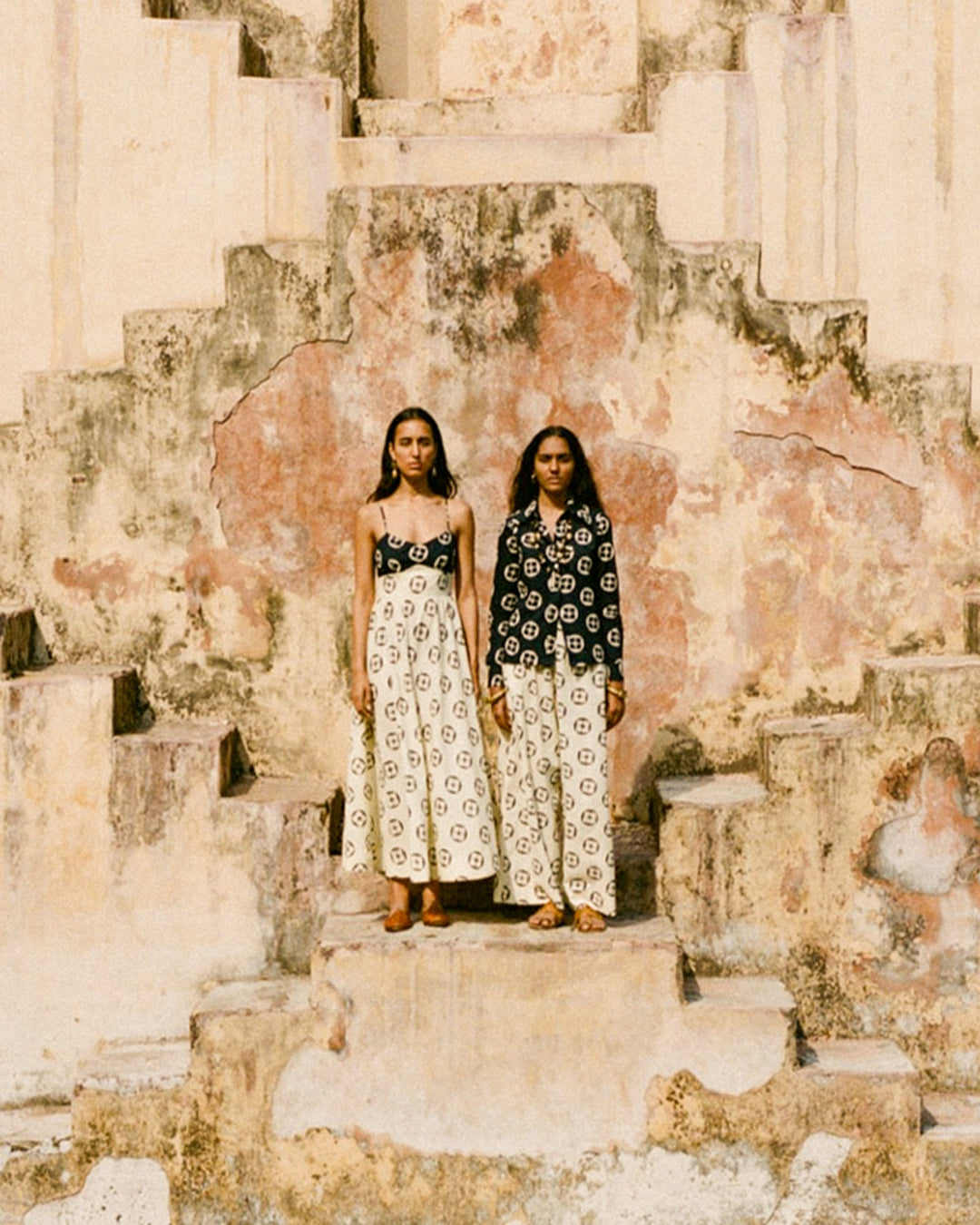 Two people stand on a weathered stone staircase, each wearing the Margie Maui Pant, known for its traditional craftsmanship and unique patterns, paired with different tops. The hand-printed cotton pants showcase elastic waists, framed by a backdrop of aged, textured walls. The earthy colors create a rustic and artistic atmosphere.