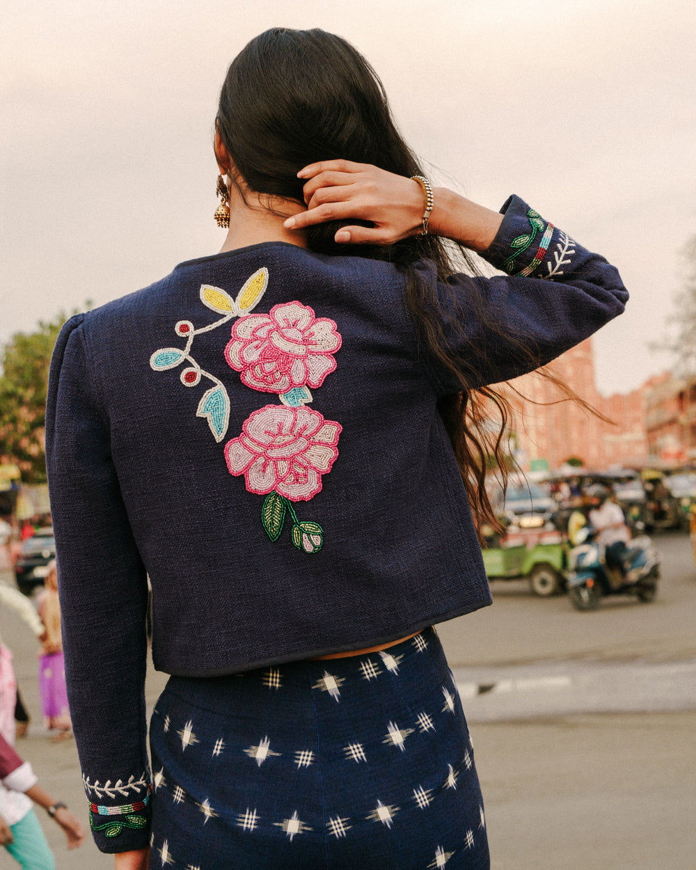 A person with long hair wears the Stella Swallow Rose Jacket, featuring vibrant, hand-embroidered floral designs on the back. They stand outdoors on a bustling street, with blurry traffic and buildings in the background.