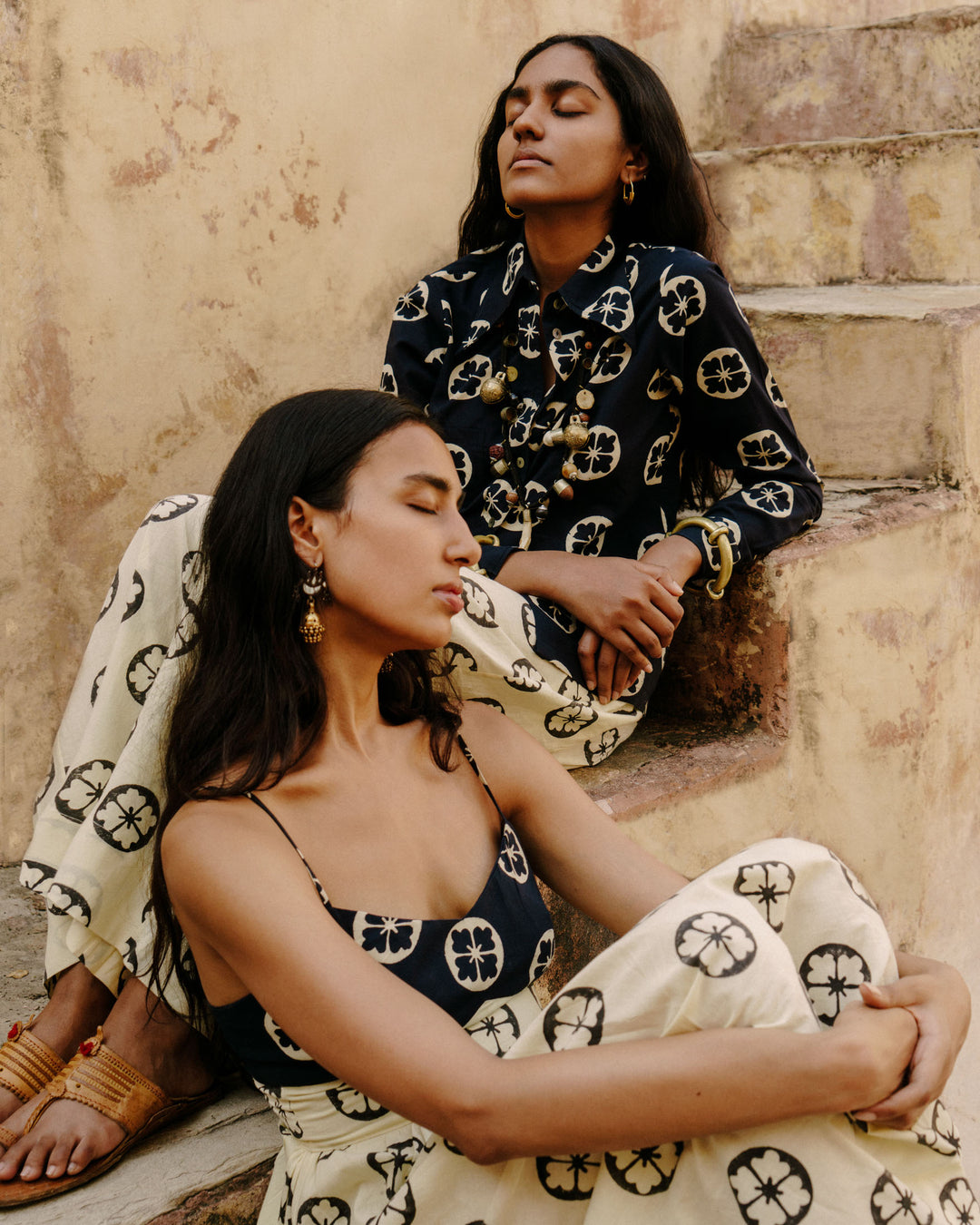 Two women sit on steps with their eyes closed, exuding tranquility in matching Alessia Callalily dresses. The smocked panel adds a touch of elegance to the navy and cream hues, while the rustic, earthy tones of the background enhance the serene and harmonious atmosphere of hand-printed floral cotton.