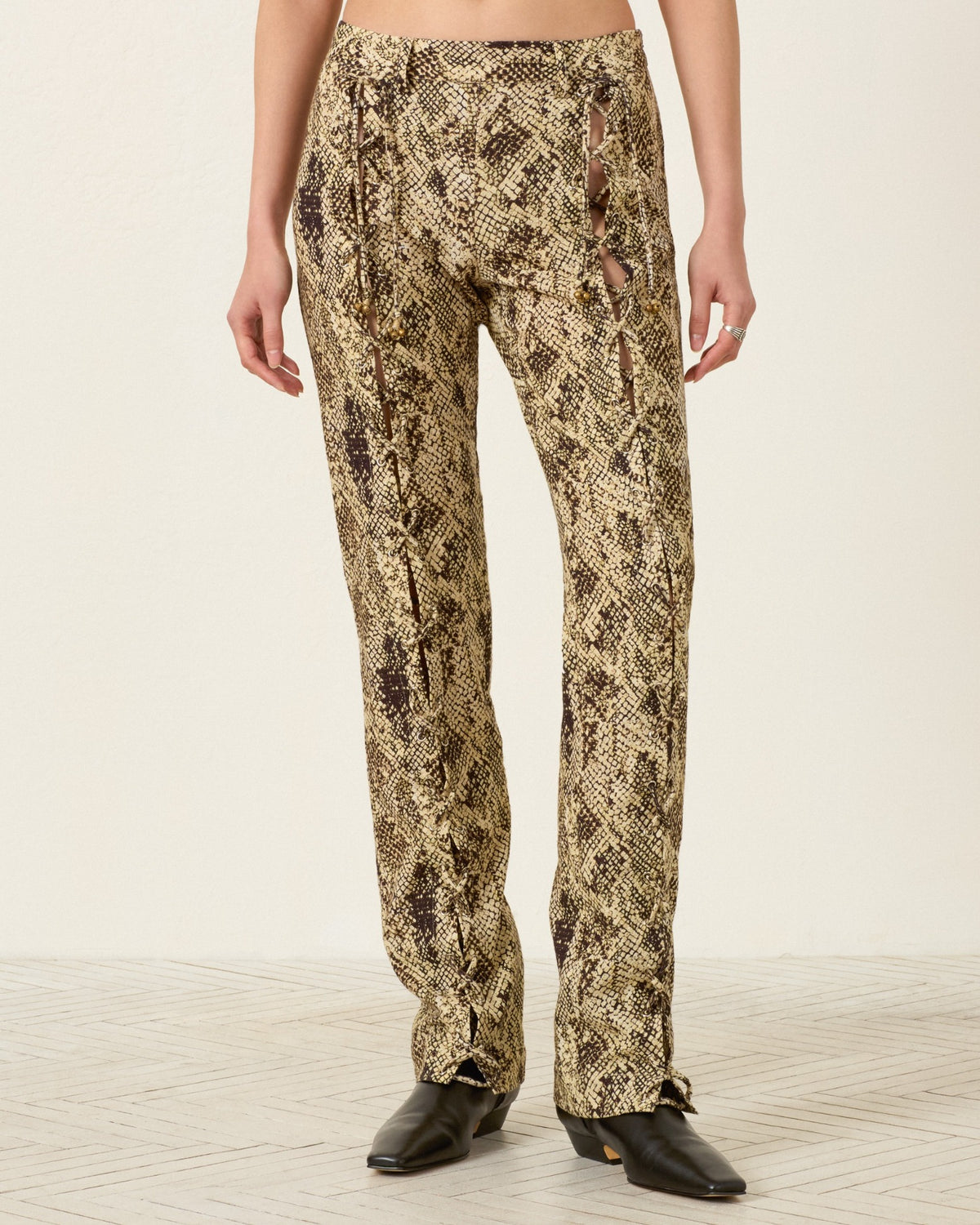 A person is wearing the Razza Lemon Snakeskin Pant, featuring a slim fit, snakeskin print, side cutouts, and lace-up ties, paired with black shoes and standing against a plain background.