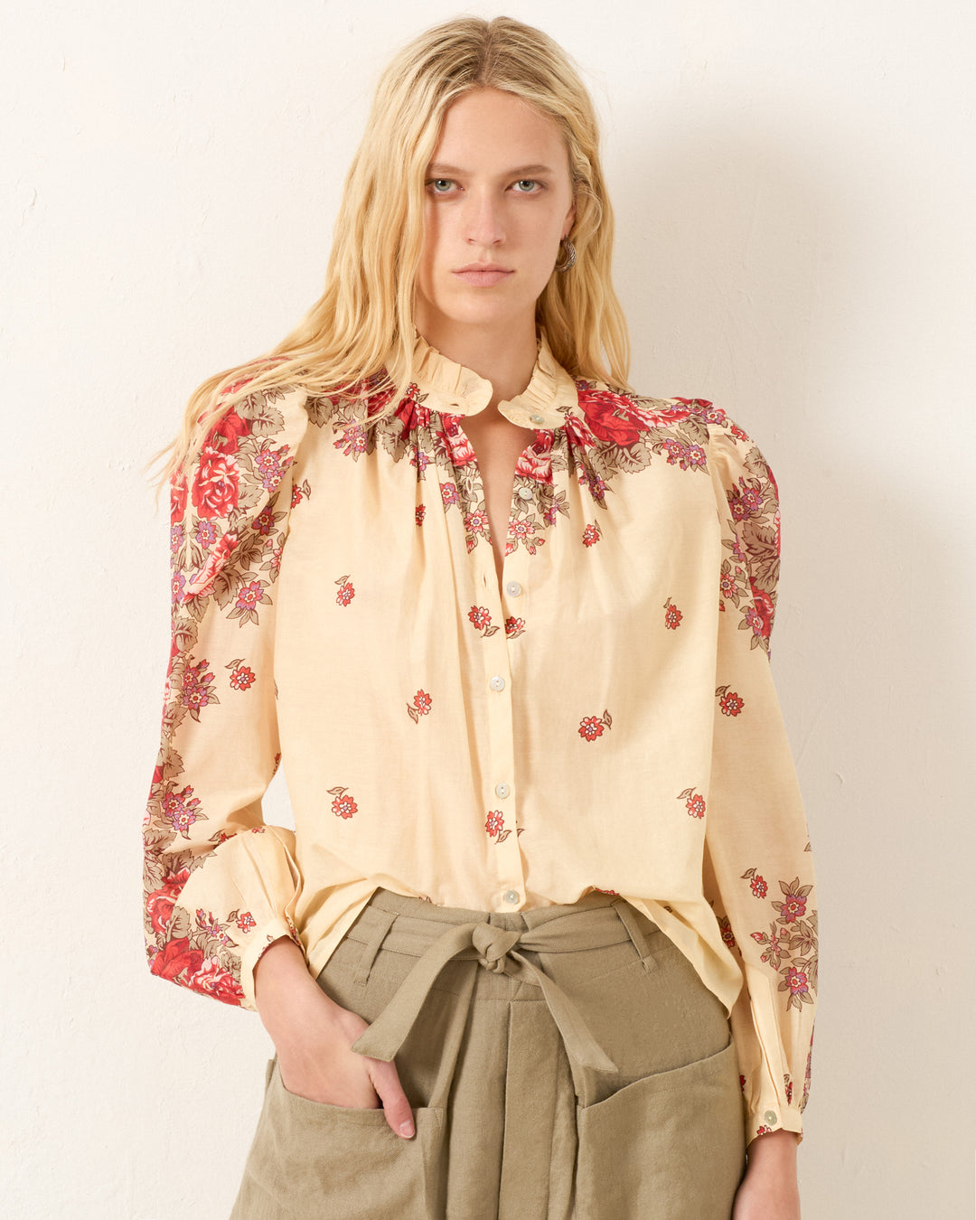 A woman with long blonde hair wears the Annabel Rosebush Shirt, a cream cotton-silk blend with red floral patterns and puff shoulders, paired with olive green high-waisted pants against a plain light background.