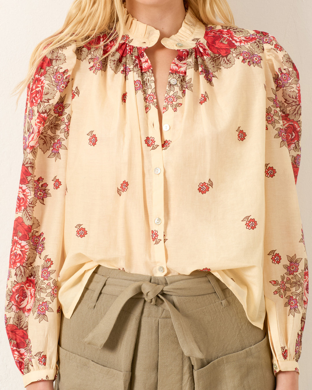 A woman wears the Annabel Rosebush Shirt, a cream cotton-silk blend with red floral print, ruffled collar, and puff shoulders, paired with high-waisted olive green pants.
