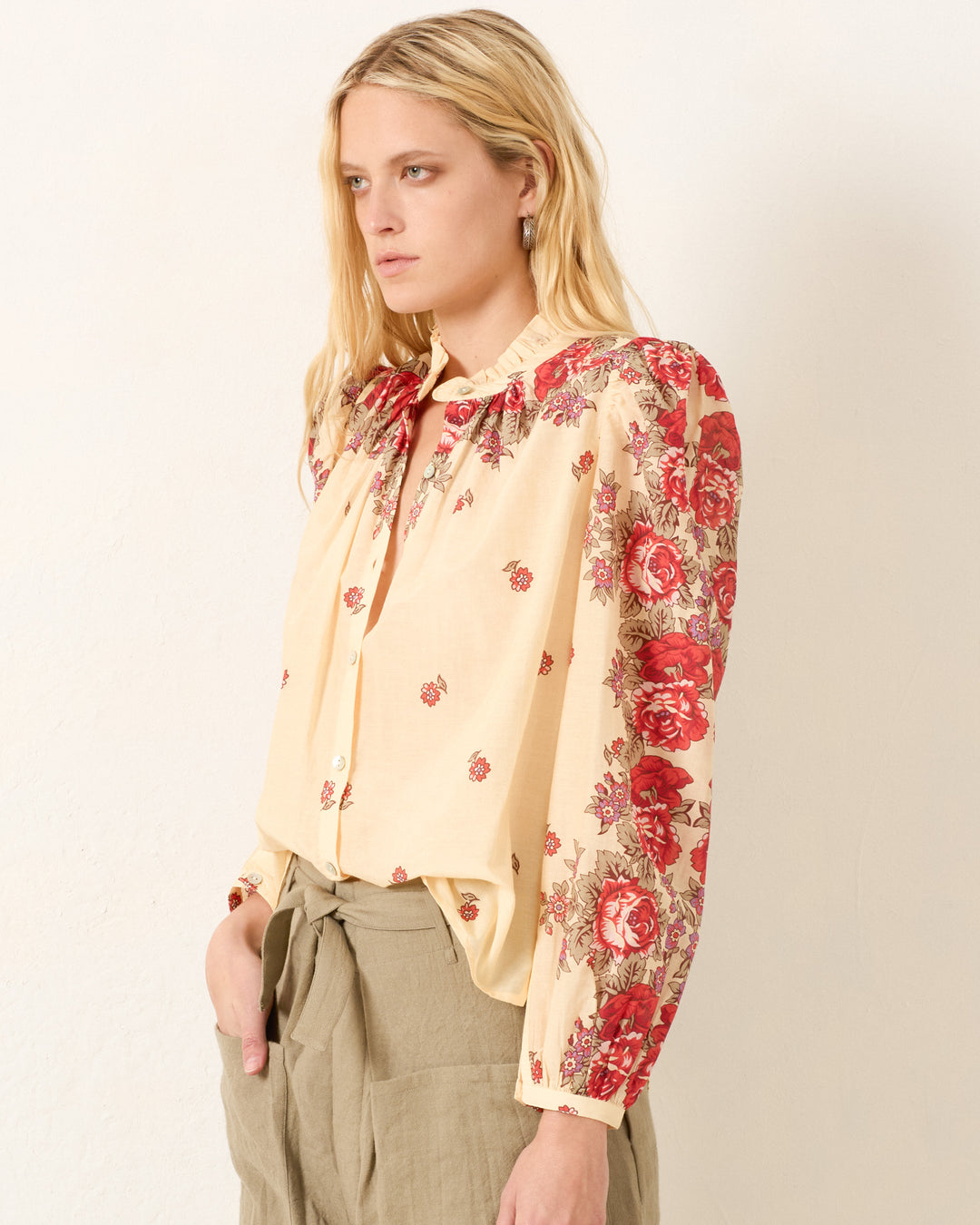 Wearing the Annabel Rosebush Shirt—a cream cotton-silk blend with red floral patterns—and beige trousers, a woman stands against a light-colored wall, gazing to the side.