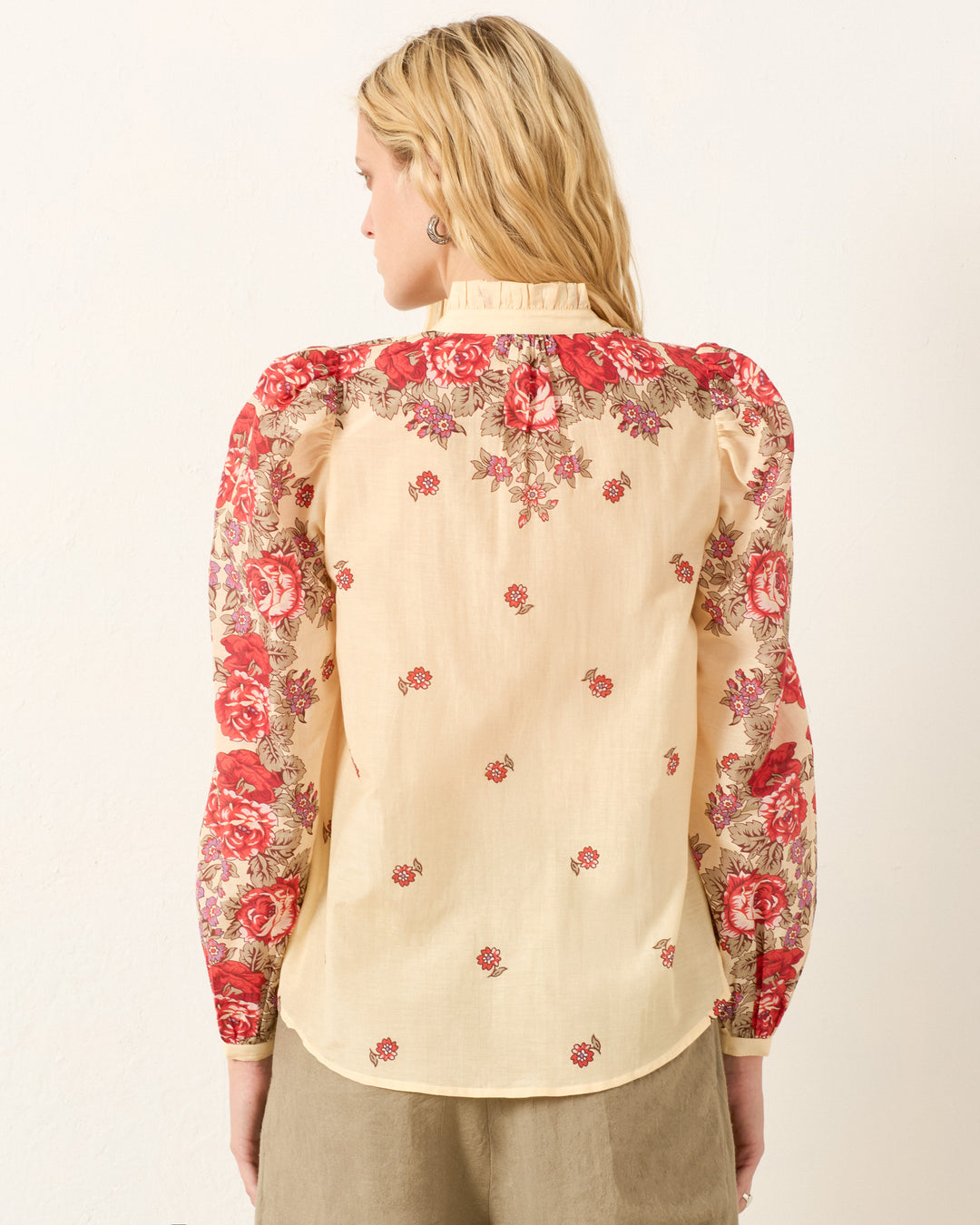 A blonde woman faces away, wearing the Annabel Rosebush Shirt—a cream cotton-silk blend with red floral patterns on the shoulders and sleeves, and a high ruffled collar.