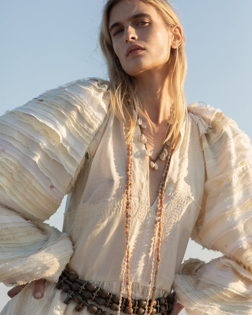 A person with long blonde hair stands outdoors under the clear blue sky, wearing the Celeste Sahara Sari Dress—a textured white dress featuring kantha quilting and layered sleeves—accessorized with long necklaces and a patterned belt.