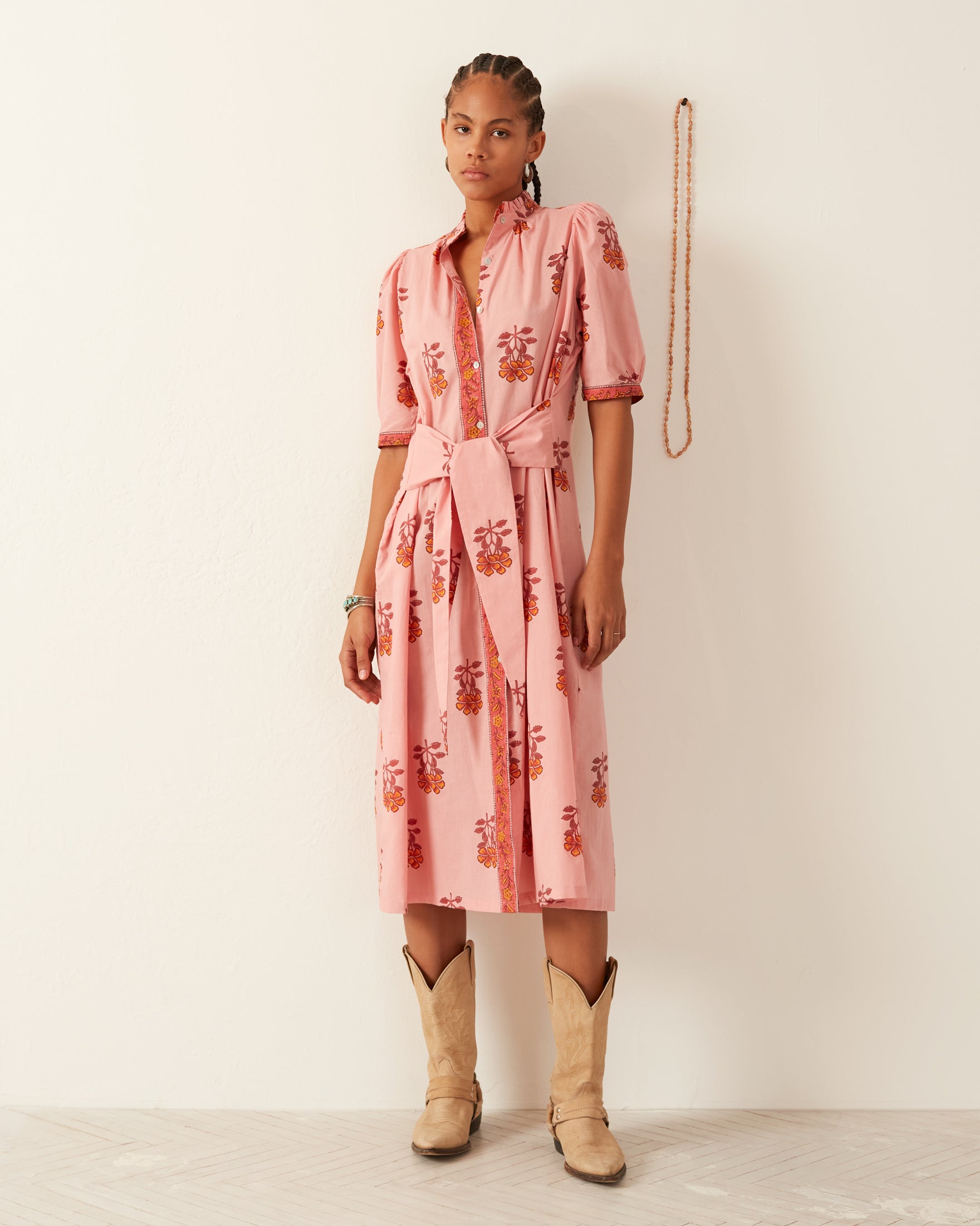 A person wearing a hand-printed cotton Remi Peony Dress, featuring a tie belt and ruffled collar, stands against a light wall. Their braided hair complements their tan cowboy boots, while a beaded necklace hangs on the wall beside them.