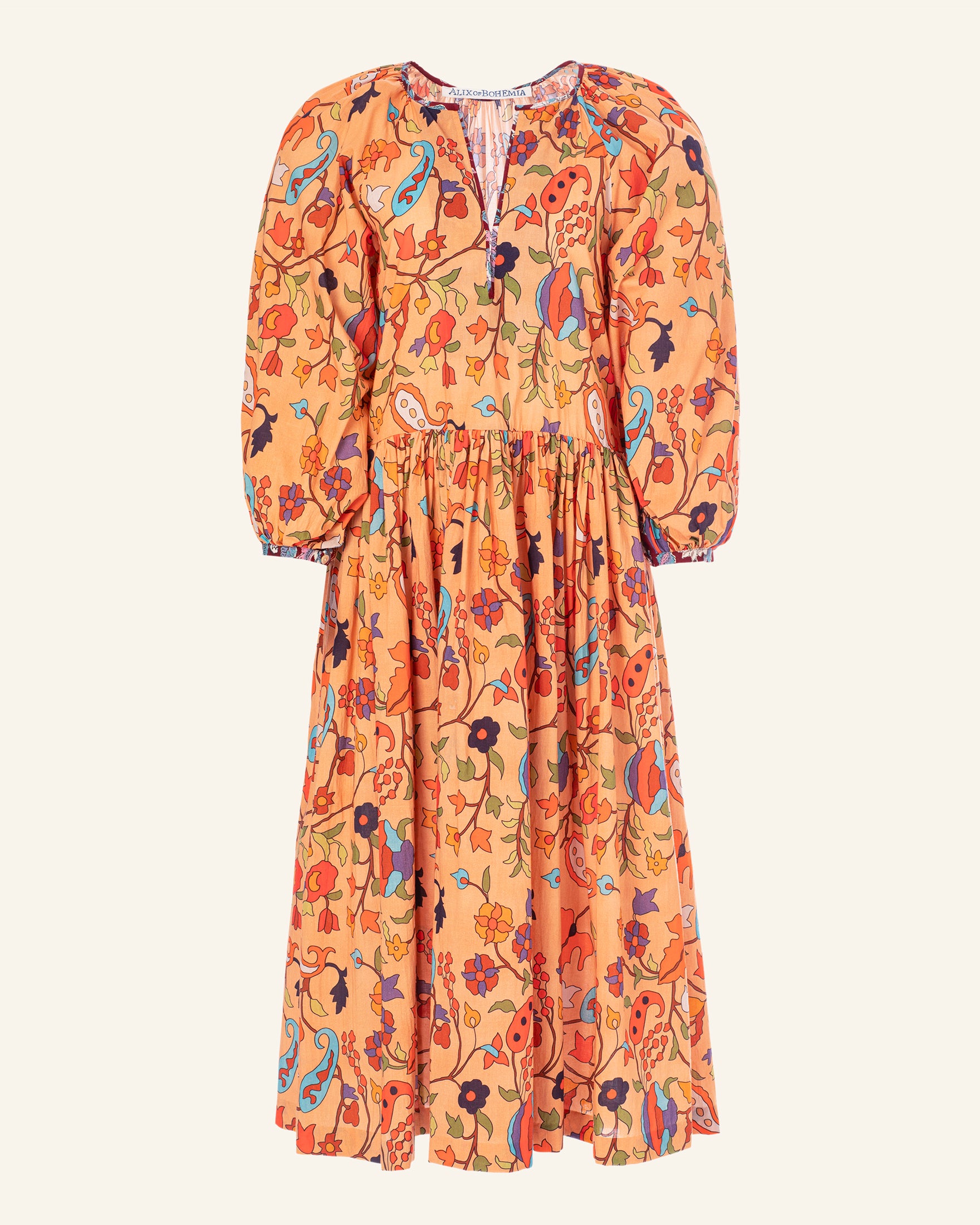 The Tuesday Tigerlily Dress is an orange long-sleeve silk cotton ensemble adorned with a floral and paisley pattern in red, blue, and green hues. It features a gathered waist and a round neckline against a neutral backdrop, highlighting exquisite hand-printed details.