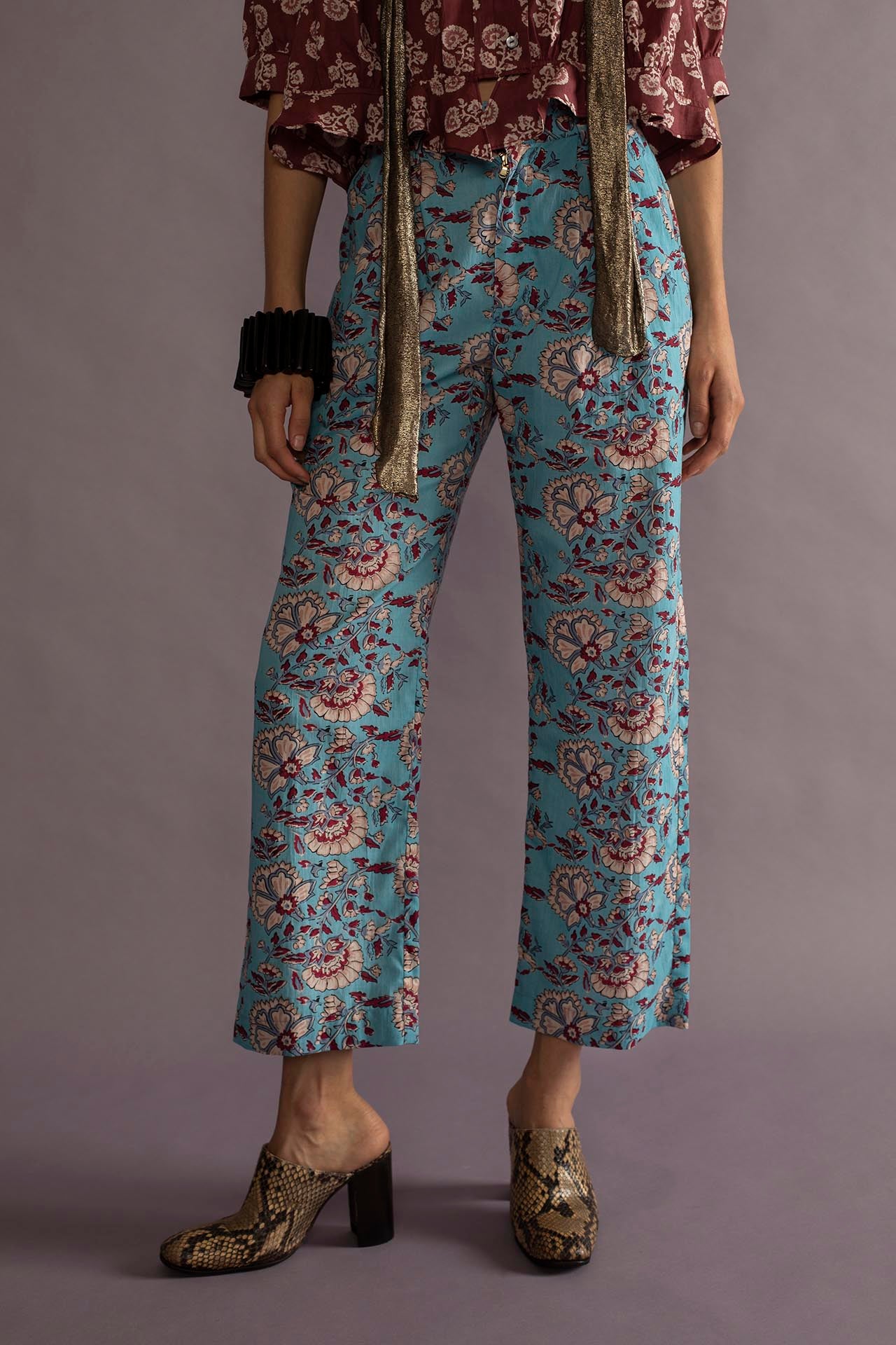 Against a gray background, a person wears a floral maroon blouse and scarf, paired with the artisan-crafted Catch Azur Pant. They complete their look with snake-print heeled shoes and a black bracelet, hands by their sides, showcasing their unique style.