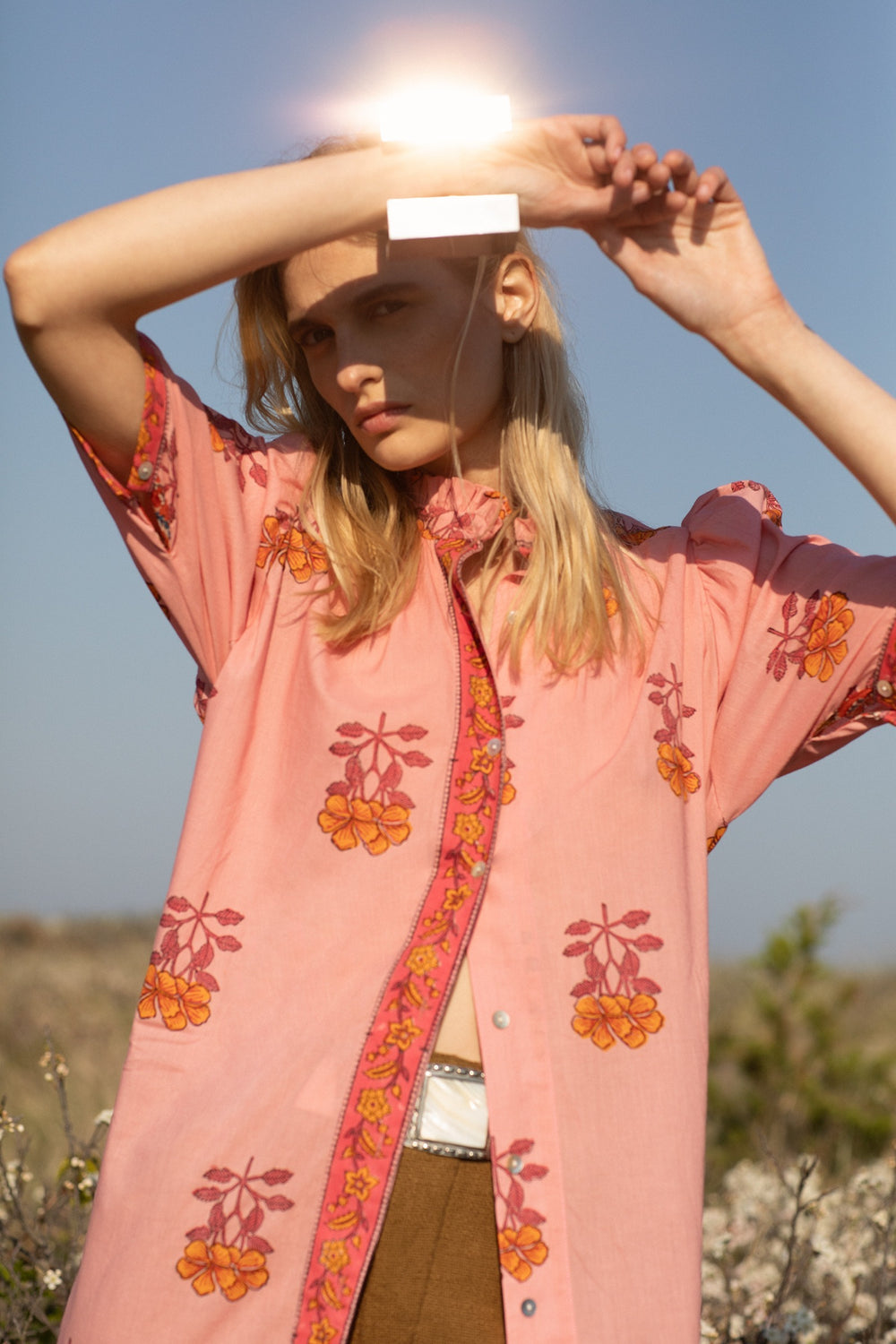 Under a bright sky, a person stands outdoors, shading their eyes with one hand. They are dressed in a pink shirt featuring a floral pattern and orange details that echo the vibrant style of the Remi Peony Dress, paired with light brown pants. The background is a soft-focus natural landscape.