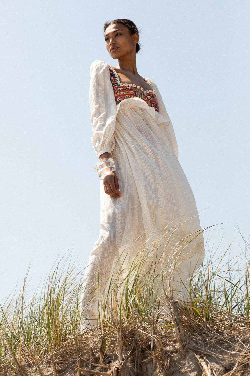 Wearing the Bibi Desert Bride Dress, a person stands in a grassy area, gazing to the side under a clear sky. The long, hand-embellished white dress features colorful embroidery near the neckline and is complemented by bracelets on one wrist. The upward perspective makes the figure appear tall and majestic.