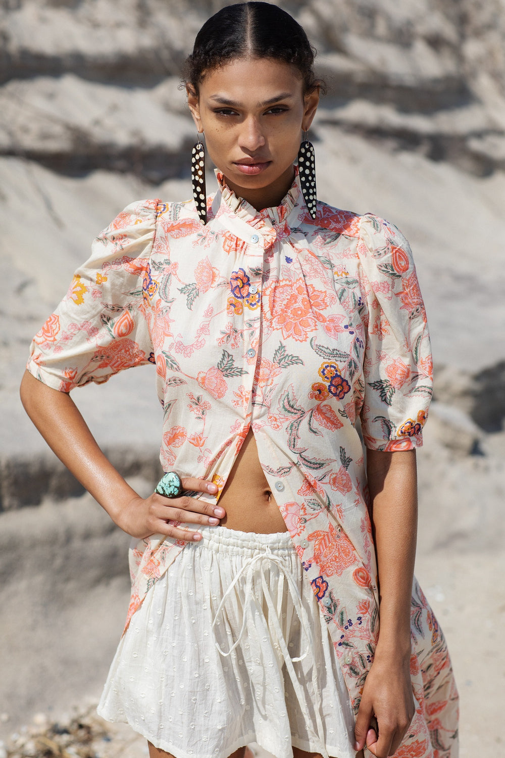 A person stands on a sandy beach, wearing a light, floral-patterned top paired with the Christy Powder Spot Short, known for its relaxed fit and elasticated waistband. They are accessorized with long, black geometric earrings and a green ring, while the ocean waves and rocks form a blurred backdrop.