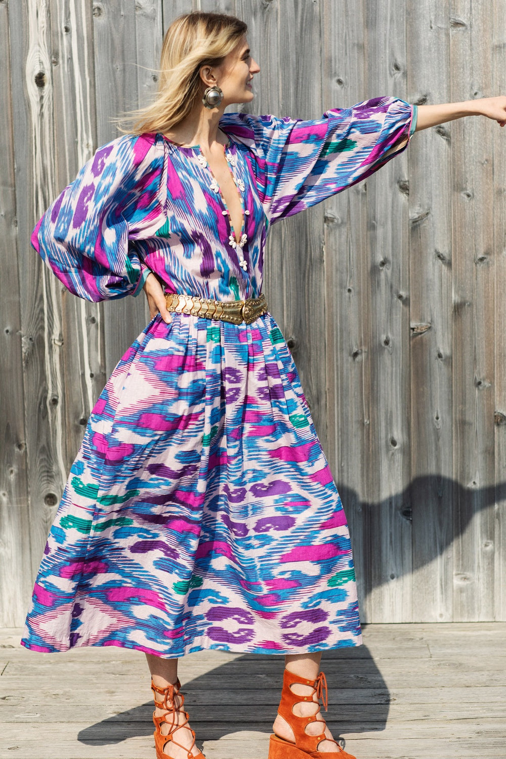 A woman wearing the Celeste Oasis Ikat Dress made of colorful, hand-printed cotton with long sleeves stands against a wooden backdrop. Her dress features vibrant pink, blue, and purple patterns created using traditional techniques. She points to the right while wearing orange lace-up sandals and large earrings.