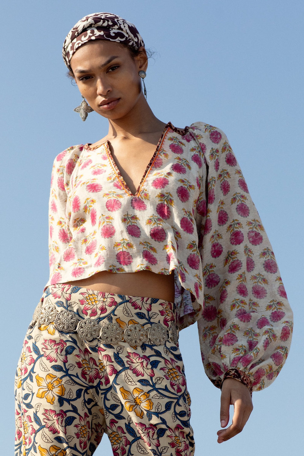 A woman poses against a clear blue sky, wearing the Marlowe Pomegranate Top with long sleeves and a matching headband. Her patterned pants boast floral designs crafted using traditional methods. Silver earrings and a detailed belt accentuate her ensemble, adding charm to the hand embroidery.