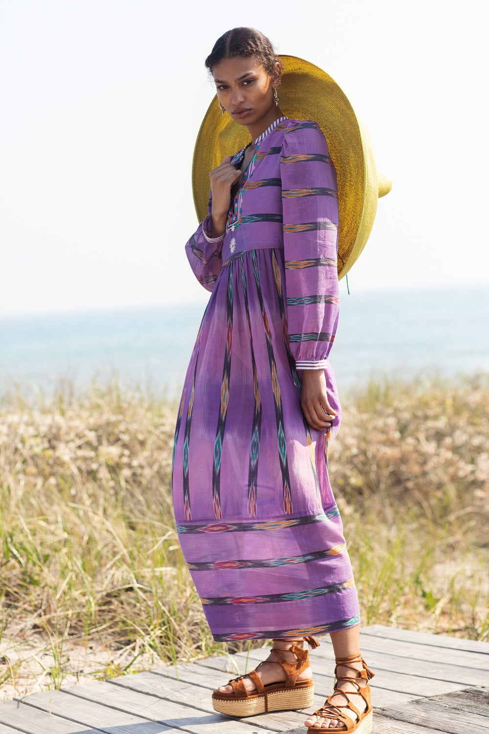 A woman stands on a boardwalk wearing the Winifred Violet Night Dress, a hand-embroidered, long patterned maxi dress, paired with a wide-brimmed yellow hat and platform sandals. Behind her are grassy dunes and the sea beneath a clear sky.