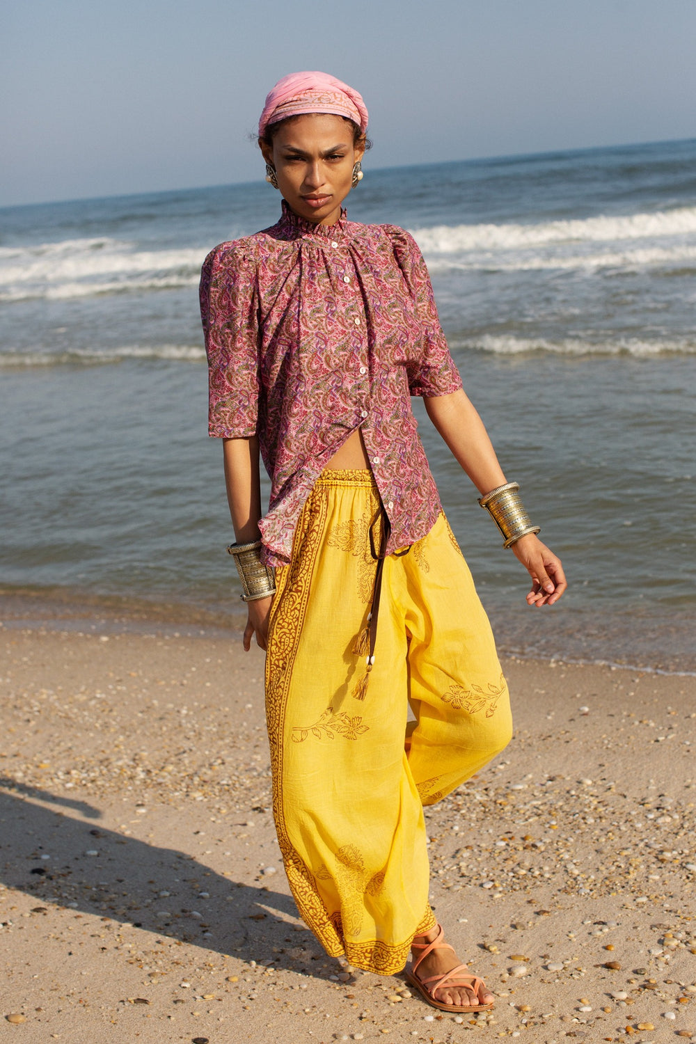 A person in a patterned pink top and Natalie Banana Flower Pant with hand block printed, wide leg yellow pants strolls along a sandy beach in sandals. They wear a pink headscarf and multiple bracelets, with the sea and blue sky as their serene backdrop.