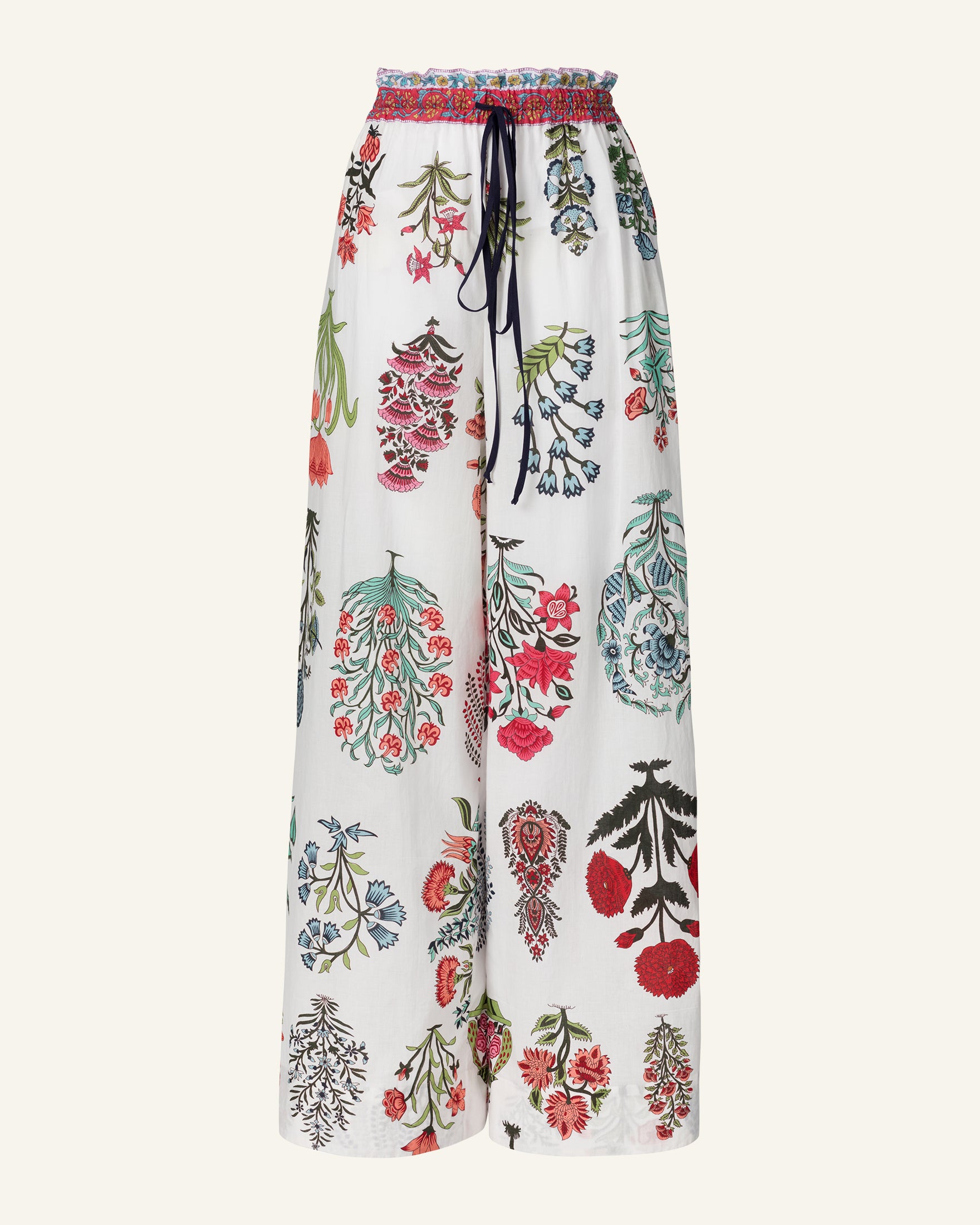 The Margie Flower Show Pant features wide-leg pants with a vibrant floral pattern of red, blue, and green blooms on hand-printed cotton. The design includes a ruched waistband and an elasticated drawstring waist for added comfort.