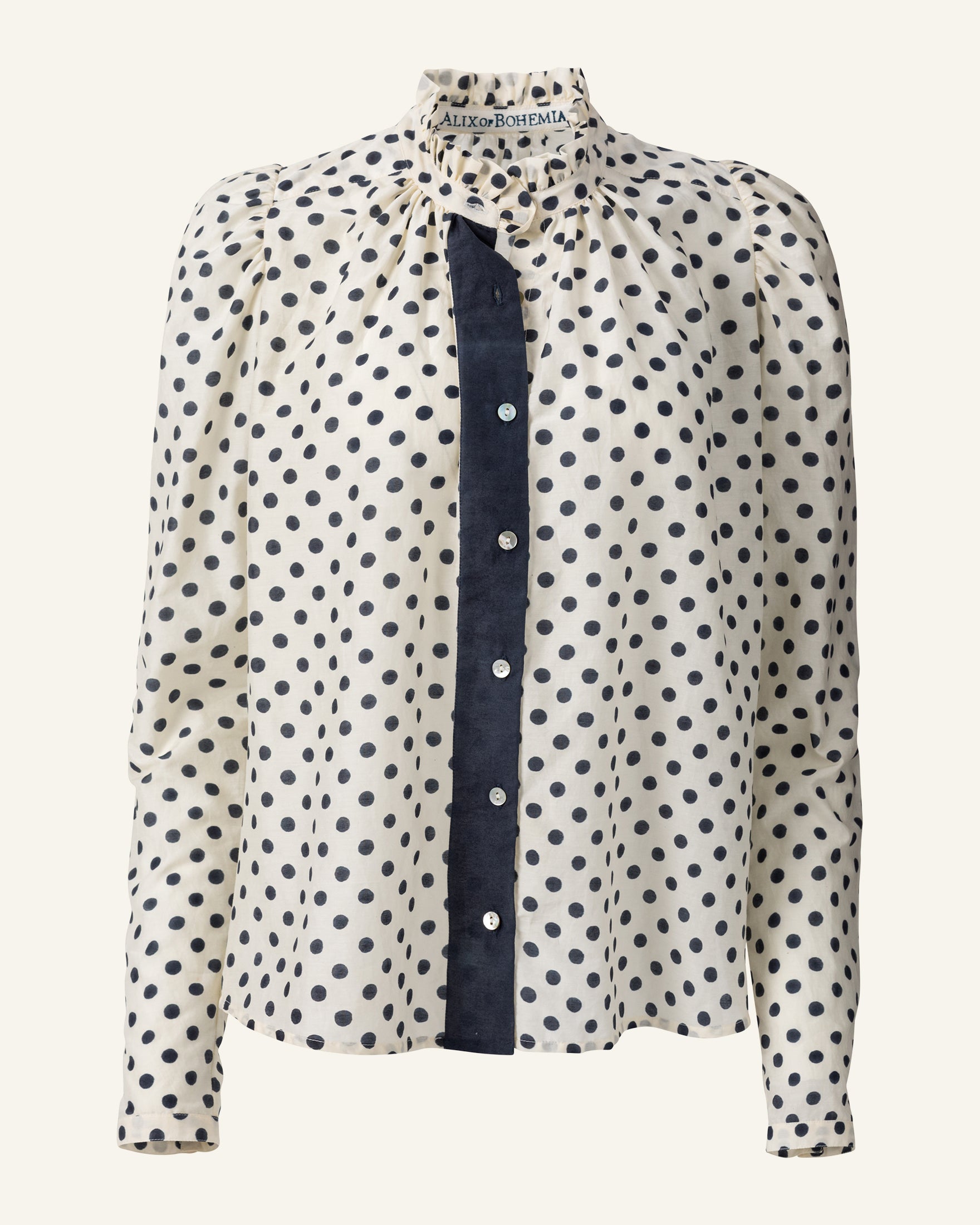 The Annabel Tuxedo Shirt is a long-sleeve blouse that boasts a high ruffled collar and puffed sleeves. It is decorated with black and white polka dots on a cream background and finished elegantly with Mother of Pearl buttons down the front.