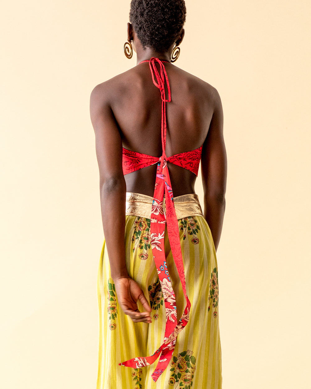 A person is seen from behind, wearing the Marmi Rosy Red Tie Top, a reversible halter style with long ties. They are also dressed in a yellow skirt adorned with floral patterns that echo traditional printing techniques. The background is a neutral beige, their hair is short, and they accessorize with large circular earrings.