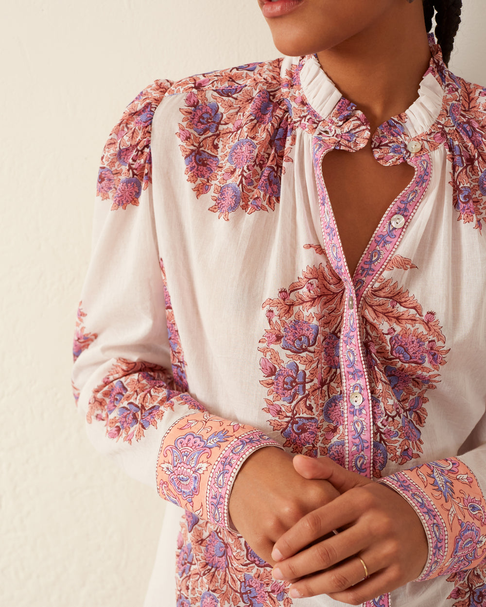 A person is wearing the Annabel Fig Flower Shirt, which showcases intricate pink and purple floral patterns on a light-colored fabric with puffed sleeves. It boasts a ruffled collar, detailed cuffs, and Mother of Pearl buttons. Hands are gently clasped in front against a neutral wall background.