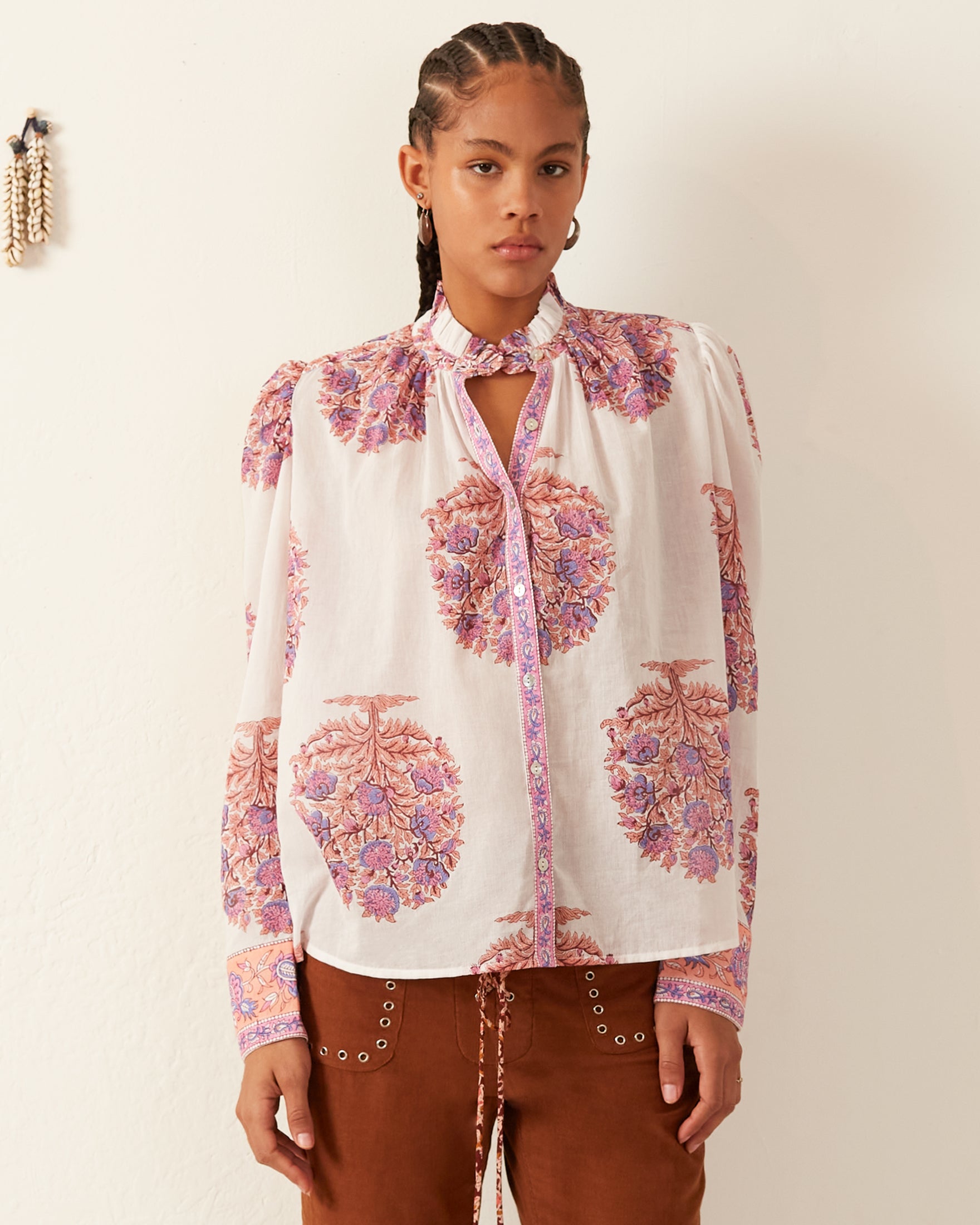 A person with braided hair stands against a white wall, wearing the Annabel Fig Flower Shirt, which features a pink and purple floral pattern, puffed sleeves, a keyhole neckline, and Mother of Pearl buttons. It's paired with brown pants adorned with white eyelet details. A decorative braid hangs on the wall.