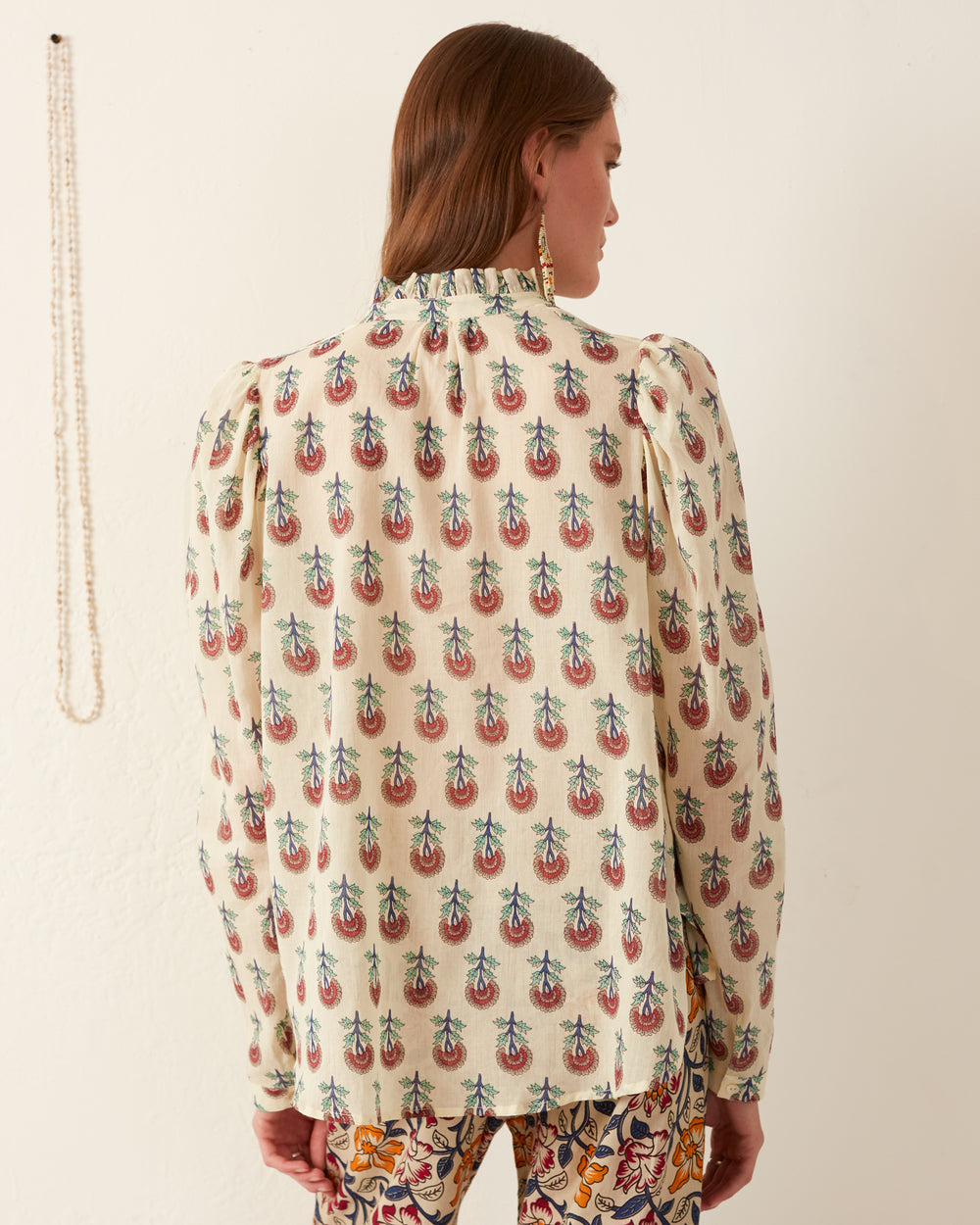 A person with long brown hair is facing away, wearing the Annabel Ivory Rose Shirt, featuring a cream hue adorned with a colorful paisley pattern and puffed sleeves. Complementing the look are patterned pants, hand block printed with intricate designs. A strand of beads hangs on the wall in the background.