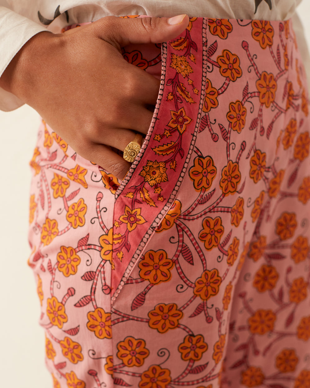 A hand rests in the pocket of the Charlie Marigold Pant, featuring pink hand-printed cotton with a vibrant floral pattern in yellow and orange. The flared leg adds a stylish touch, perfectly complementing the white top, while a ring with a floral design highlights the traditional methods of craftsmanship.