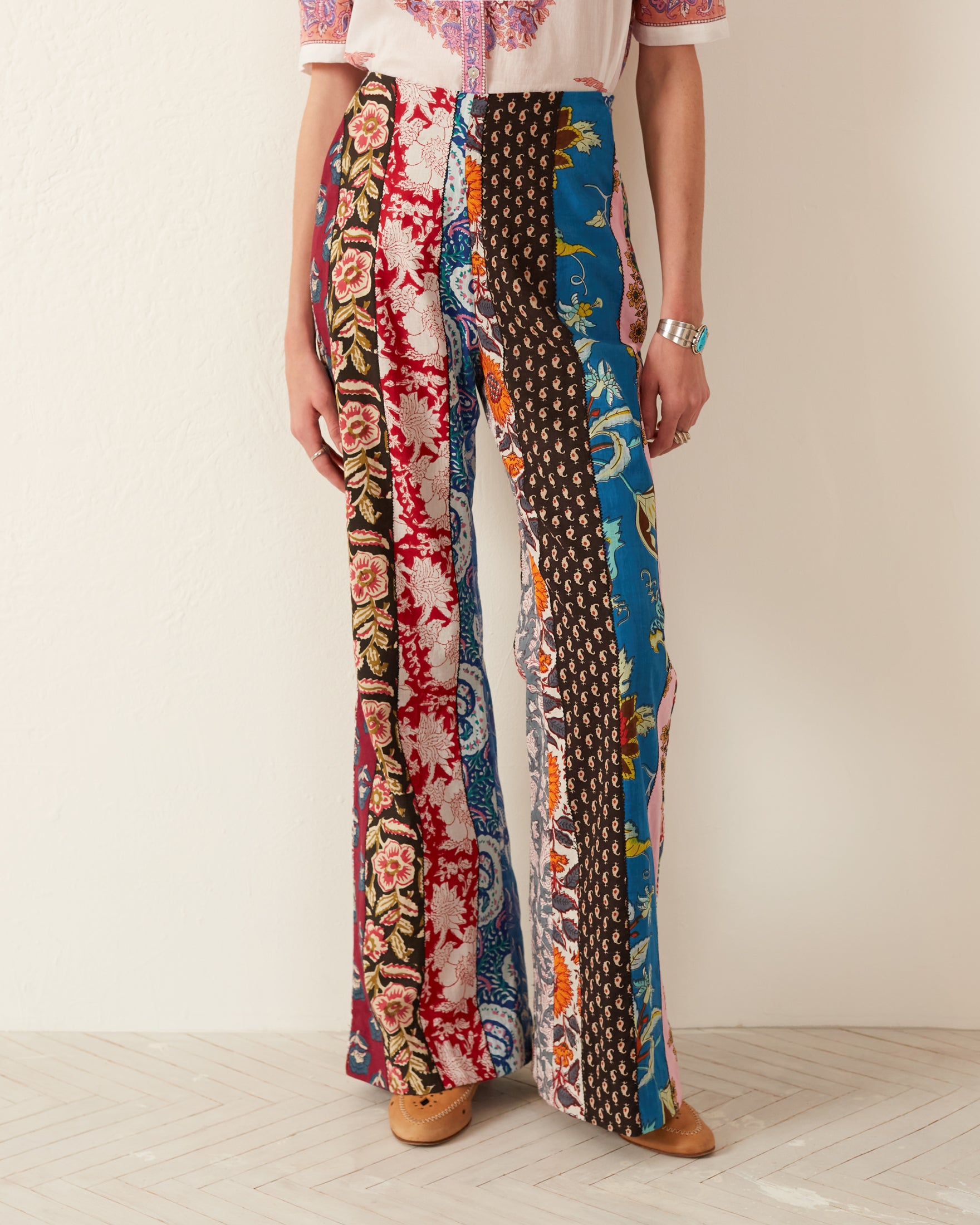 A person sporting the Charlie Wandering Patchwork Pant—a vibrant, flared-leg pair of pants made from hand-printed cotton adorned with various floral and patterned designs in bold colors—stands against a plain wall. Their ensemble is completed with a detailed, colorful blouse and tan shoes.