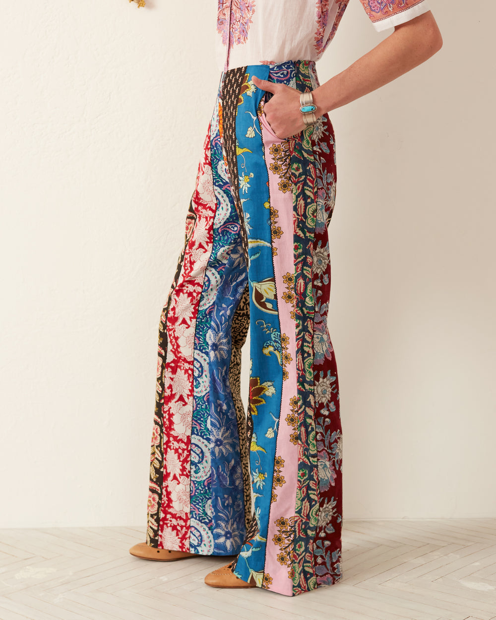 A sideways-standing individual has their hands tucked into pockets, sporting the Charlie Wandering Patchwork Pant. These vibrant pants, crafted from hand-printed cotton and featuring vertical strips of blue, red, pink, and brown floral patterns, beautifully display artisan craftsmanship. A partial view of a short-sleeved white top complements the look.