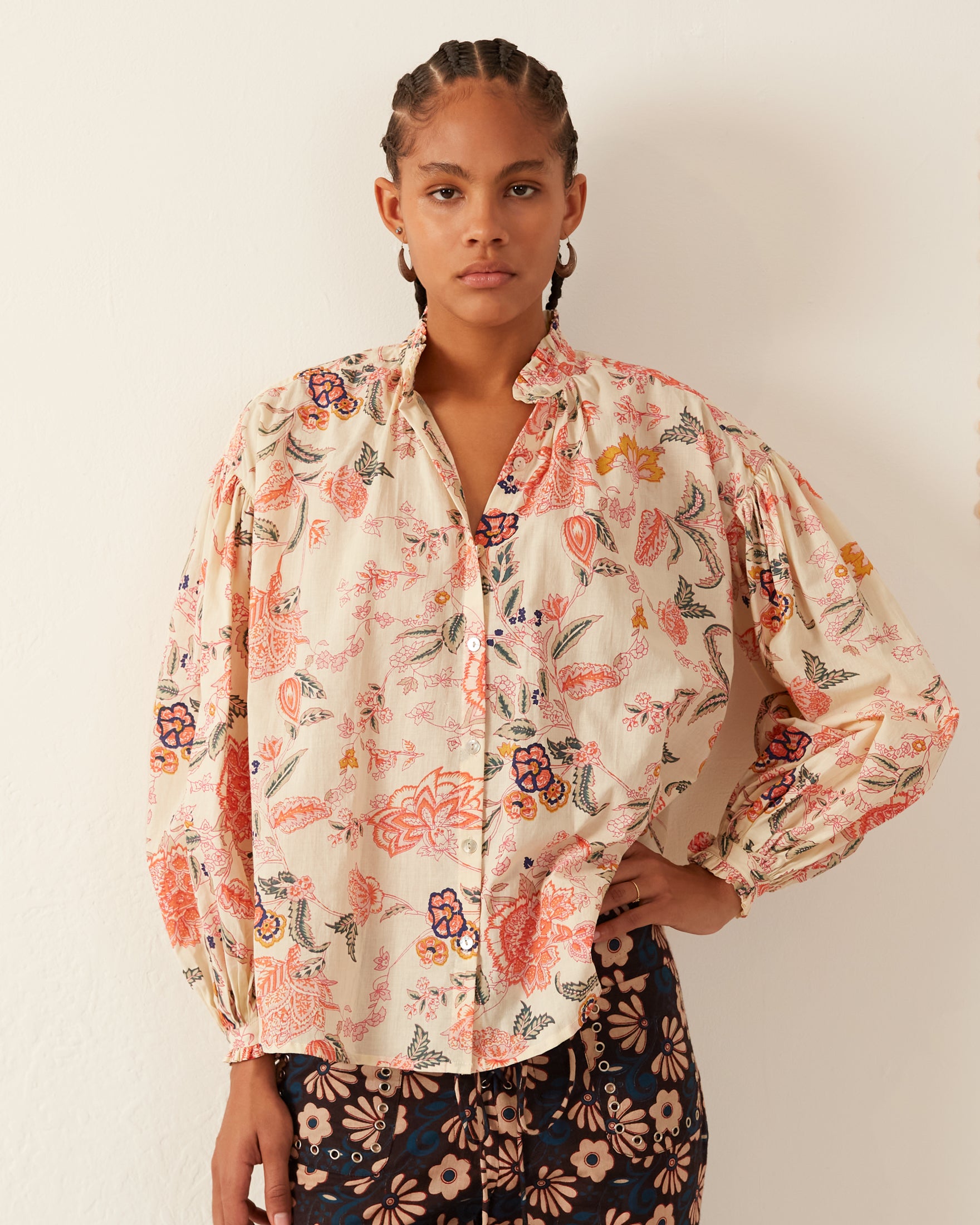 A person wearing the Poet Coral Flower Blouse, featuring a cream-colored floral design with a ruffled collar and long, puffed sleeves, pairs it with dark floral pants. Their hair is styled in braids, all crafted from hand-printed cotton as they stand against a plain white background.