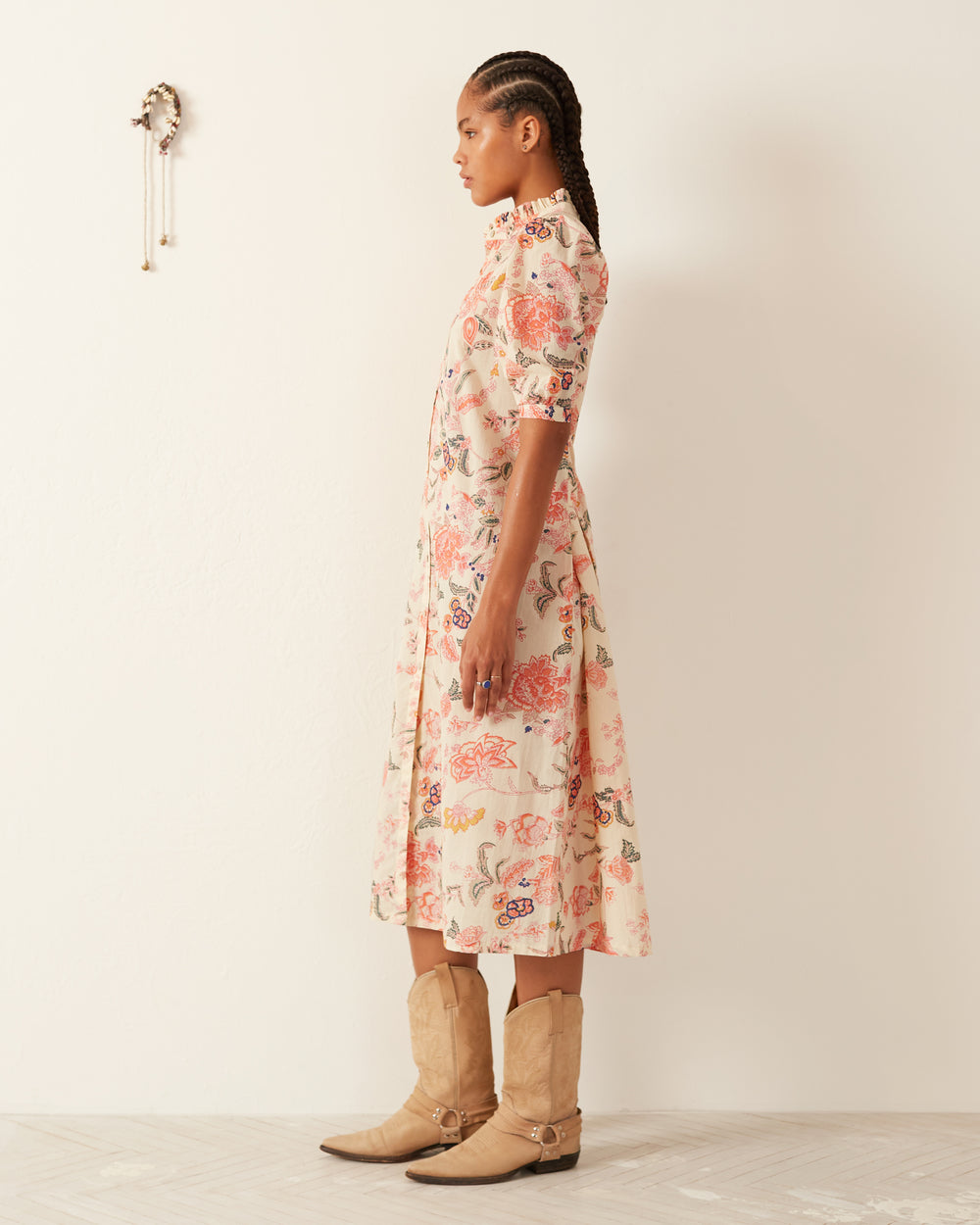 A person with braided hair stands in profile against a white wall, wearing a Remi Coral Flower Dress made of hand-printed cotton and tan cowboy boots. A small wall hanging decorates the space behind them, showcasing traditional methods of artistry.