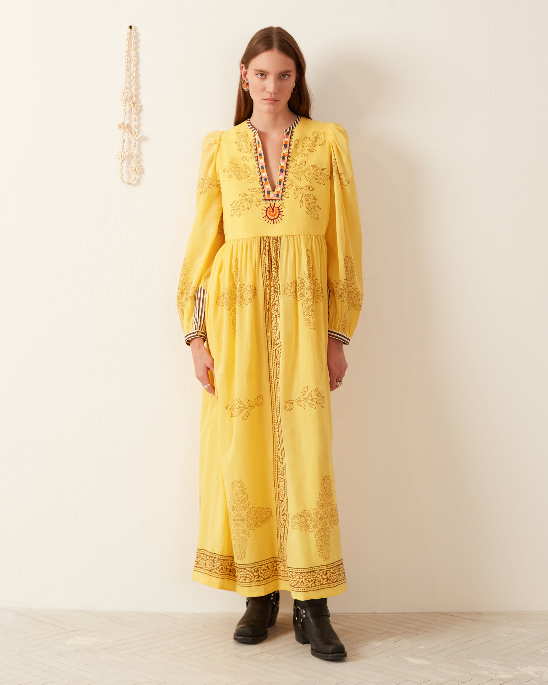 A person stands against a white wall, wearing the Winifred Amber Dust Dress, a long yellow garment adorned with intricately hand-printed cotton patterns and embellishments. They have long hair and are wearing black boots. To the left, a string of beads hangs on the wall.