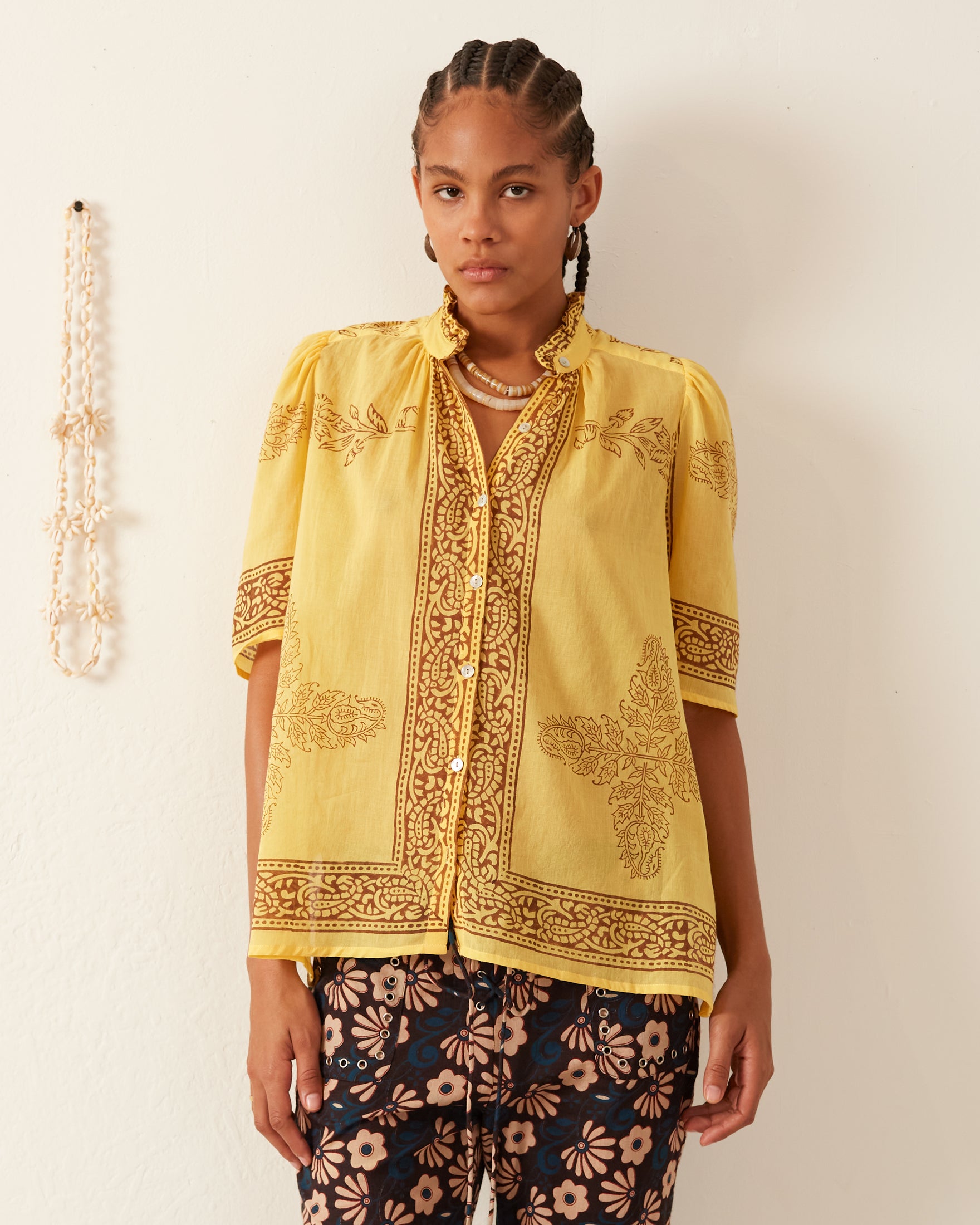 A person stands against a plain background wearing the Winnie Banana Flower Shirt, featuring a ruffled collar and paired with dark floral pants. Their hair is braided, and they maintain a neutral expression. A beaded necklace is hanging on the wall next to them.