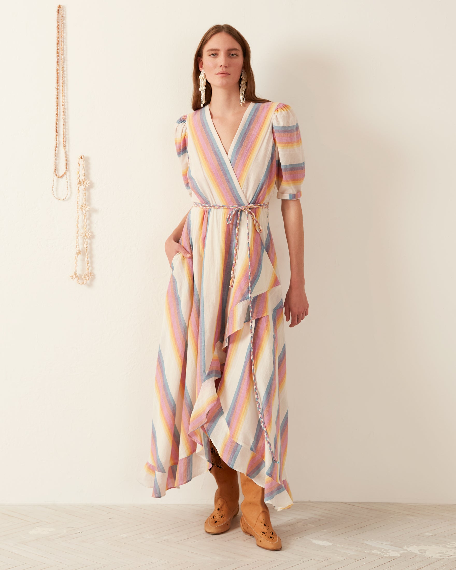 A person stands in a room wearing the vibrant Lola Sunset Wrap Dress, a colorful, striped garment made of hand-loomed cotton, featuring puff sleeves and a ruffled hem. The outfit is accented with a braided belt and paired with tan knee-high boots and large earrings. A wall adorned with hanging necklaces serves as the backdrop.