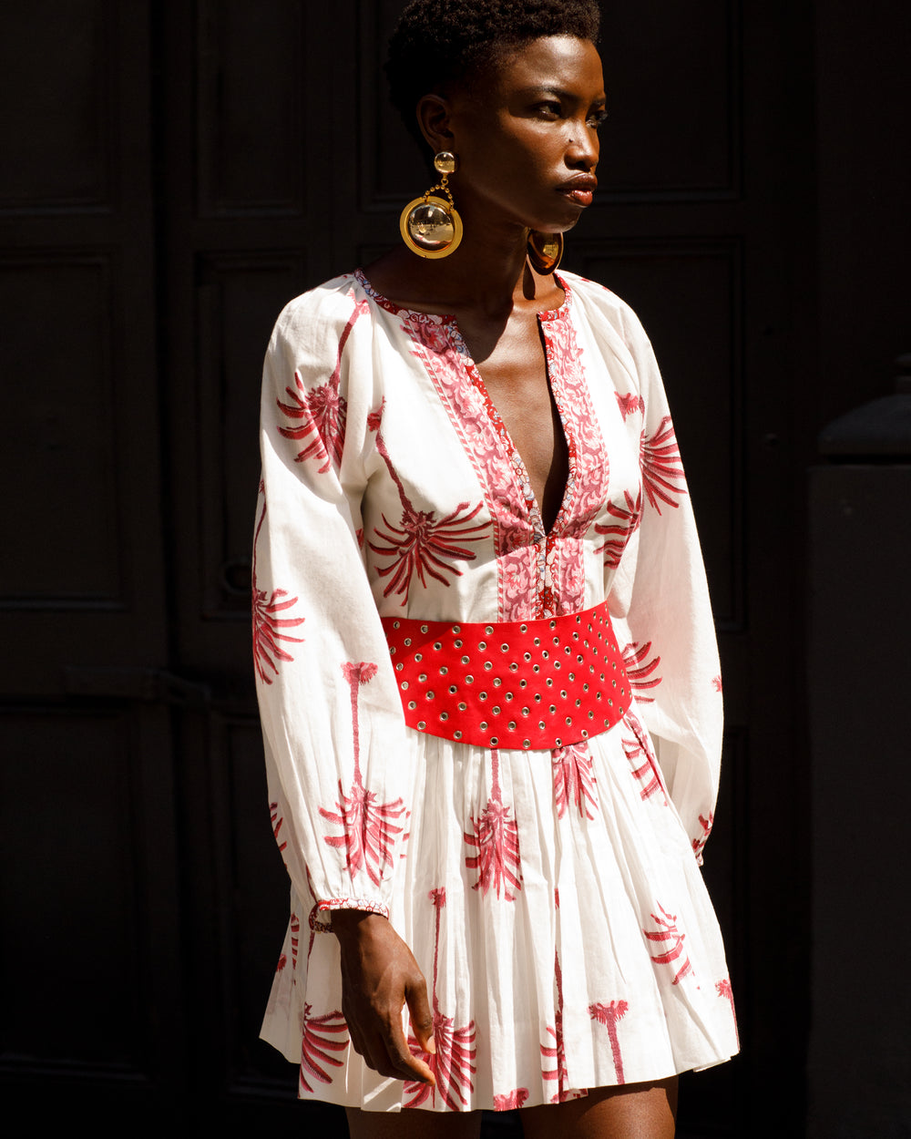 Wearing the Sula Pink Palm Dress, featuring a white design with red patterns and a gathered neckline, an individual strides against a dark backdrop. Crafted from hand-printed cotton and cinched at the waist by a red belt, they complement their ensemble with large gold hoop earrings. With short hair slightly turned to the right, they exude timeless elegance.