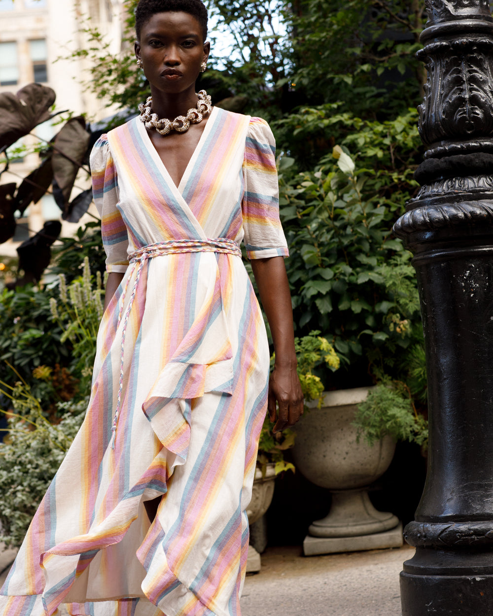 A person walks confidently in a vibrant and striped Lola Sunset Wrap Dress, crafted from hand-loomed cotton, complete with a braided belt and accessorized with a chunky necklace. They are outdoors, standing near lush greenery and a decorative light post.