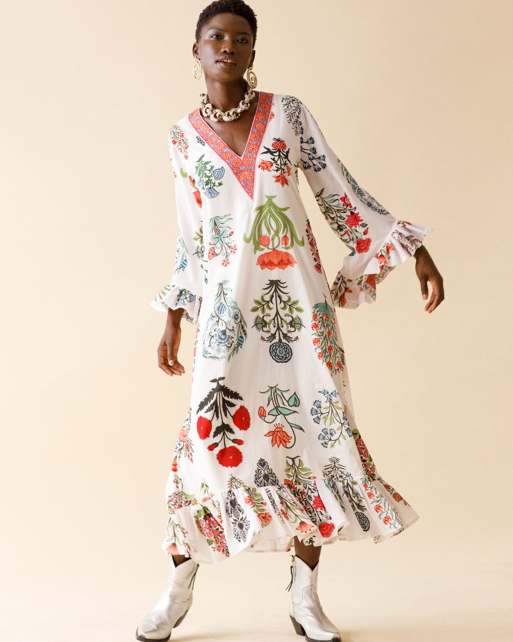 A model wearing the Zelda Flower Show Dress, meticulously crafted from hand-printed cotton, poses against a beige backdrop. This long dress showcases a white base adorned with vibrant floral patterns and includes details such as ruffled sleeves, a V-neckline, and a ruffled hem. The look is accessorized with large hoop earrings and complemented by white ankle boots.