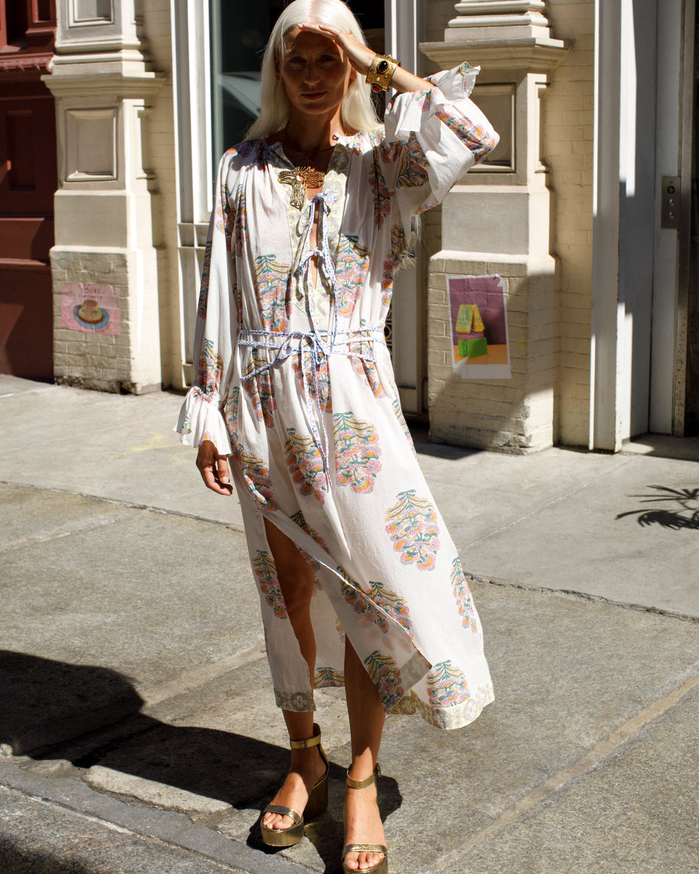 A person with long blonde hair stands on a sunlit sidewalk, wearing the Martha Honeysuckle Dress—a flowing white gown with colorful patterns hand printed on cotton using traditional methods. They wear gold sandals and are in front of a building with ornate architecture.