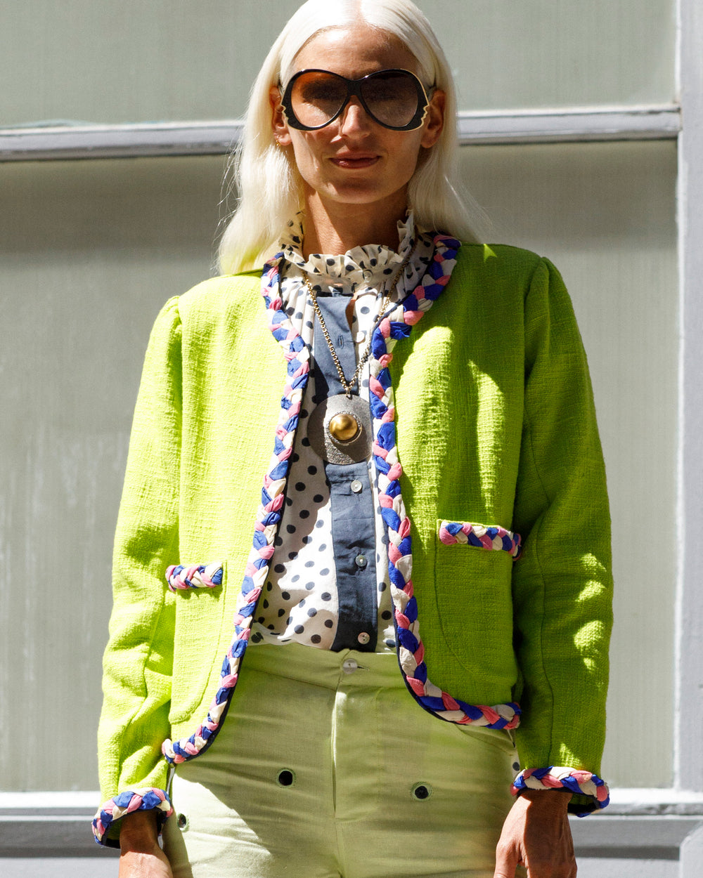 A person with long blonde hair and large sunglasses is wearing the Marilou Acid Jacket, which features handmade braided trim, along with a polka dot blouse and green pants. The jacket includes patch pockets that add flair. They are standing in front of a window, looking directly at the camera.