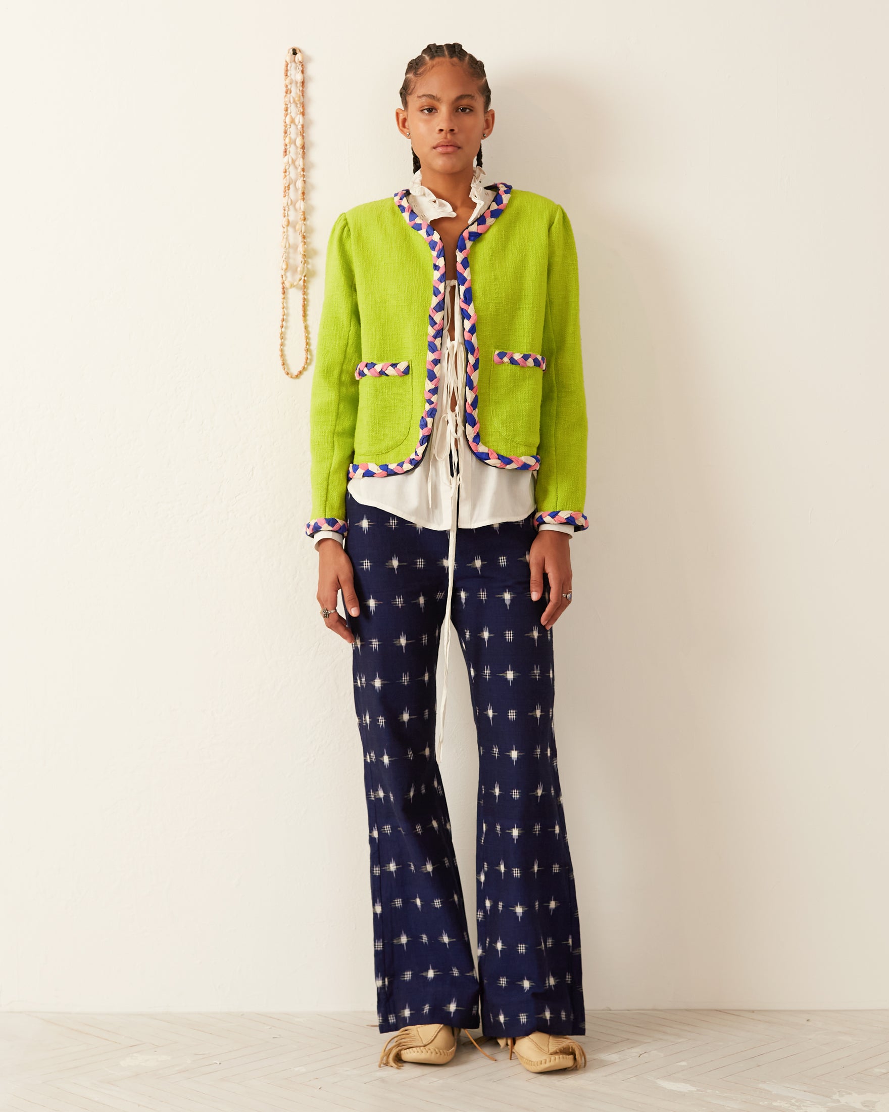 A person stands against a white wall, wearing the Marilou Acid Jacket featuring a bright green hue with handmade braided trim. They pair it with a white shirt and navy blue pants adorned with a star pattern. A decorative string embellished with beads hangs on the wall, contrasting against the light wooden flooring.